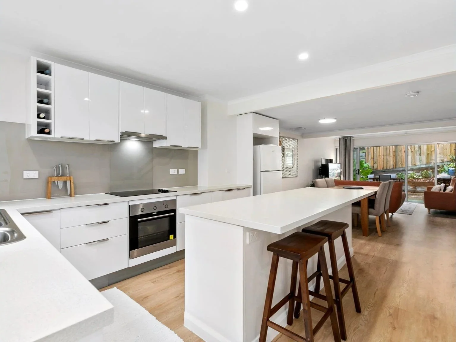 Open-plan kitchen and living room area with white cabinets, a kitchen island, wooden stools, and seating with a view of a backyard outside.