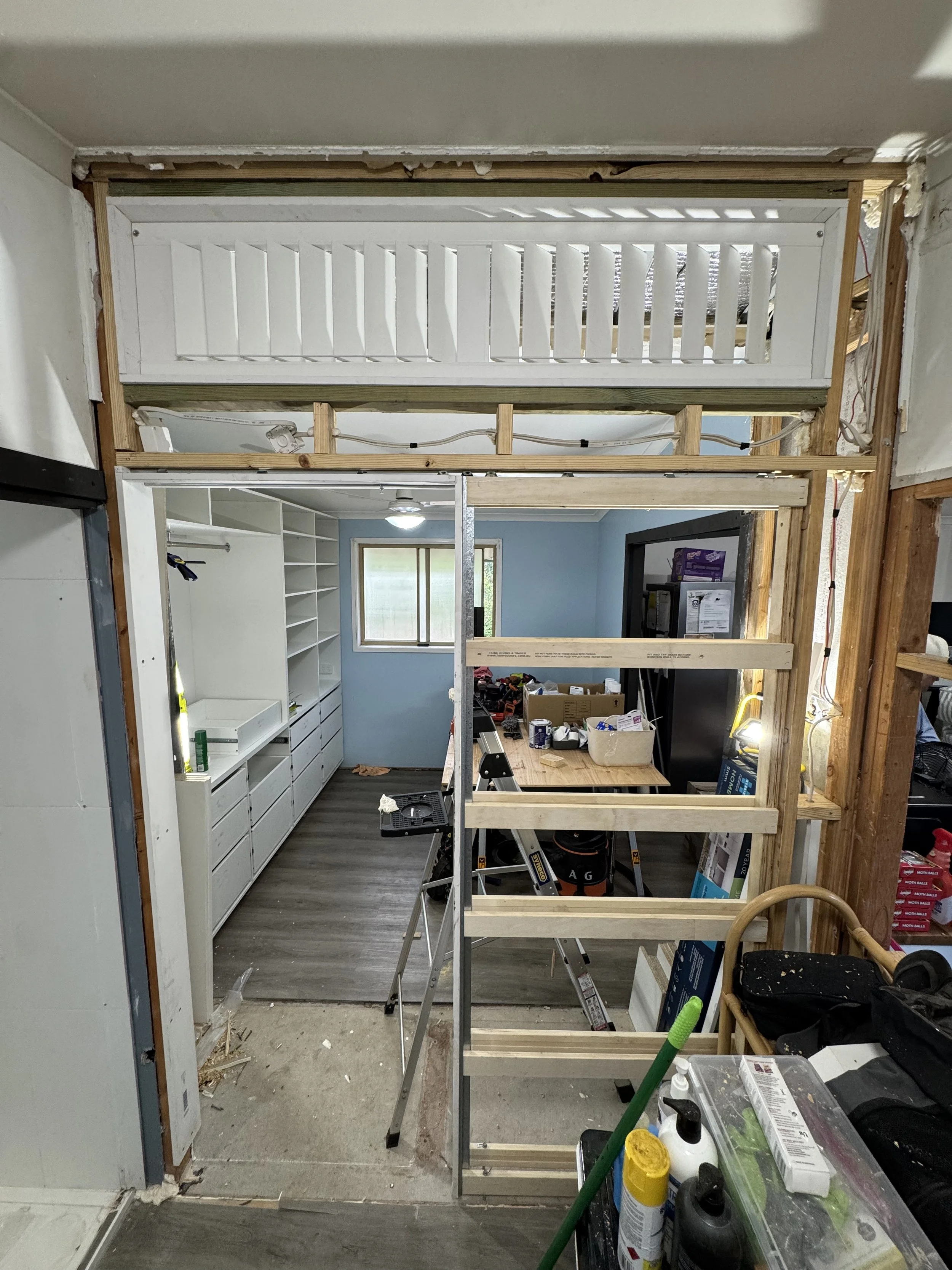 Room under renovation with framing for a new wall, partially finished walls, construction tools, supplies, and a window in the background.