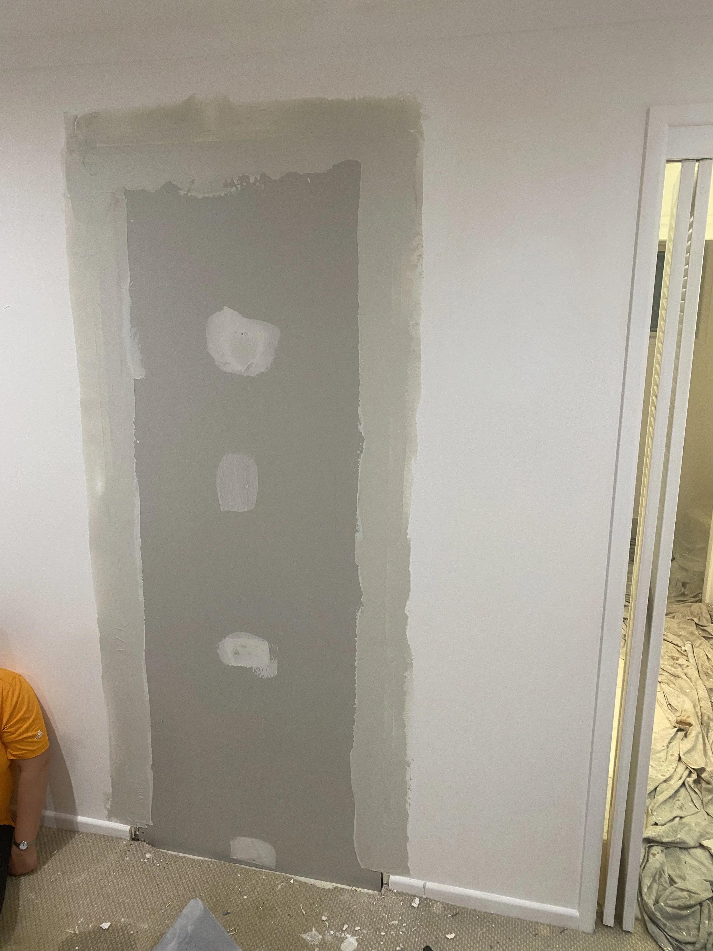 Interior wall construction with drywall installation, showing partially finished drywall with patches of joint compound and tape, and an open doorway on the right side.