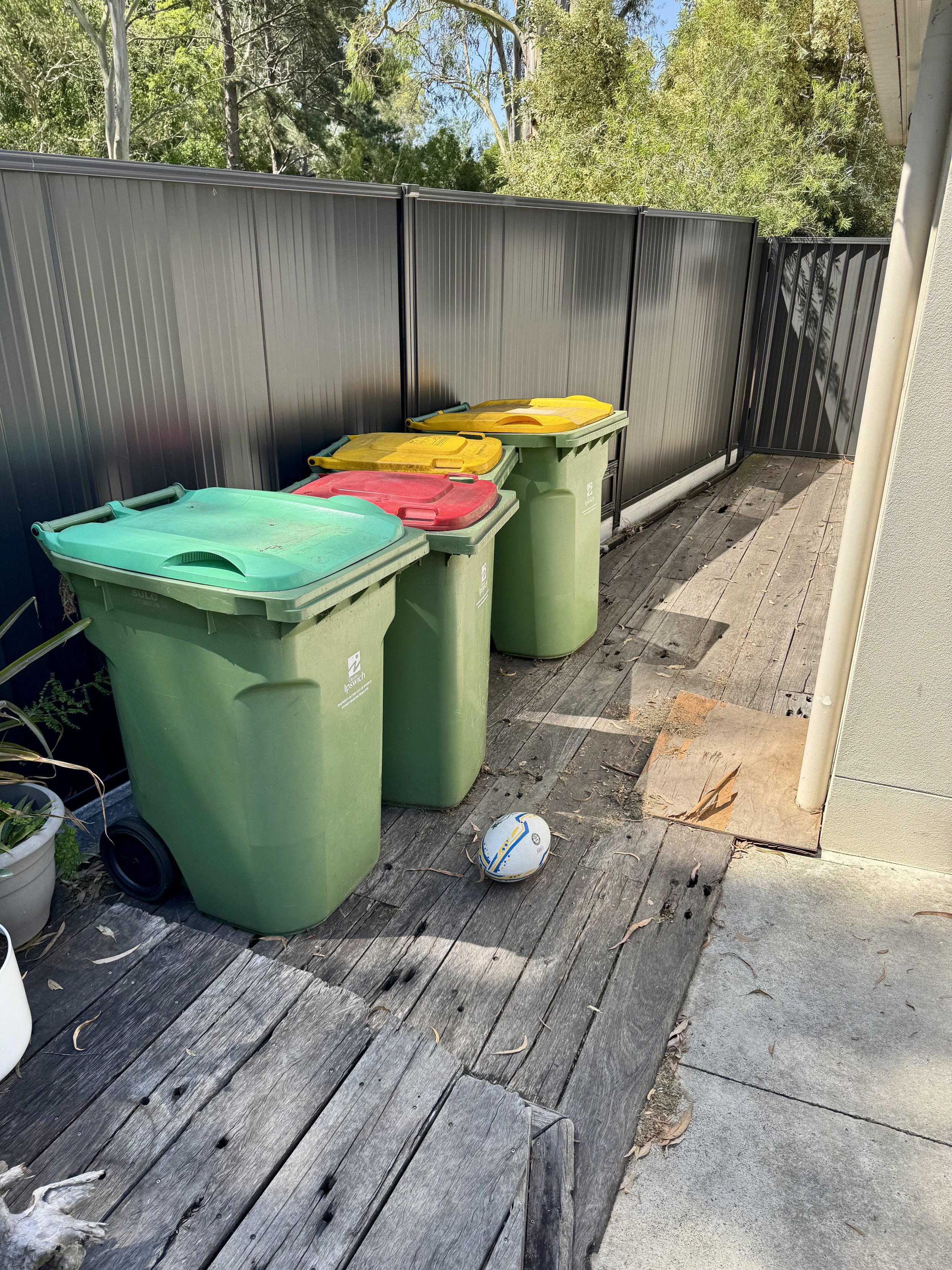 Four garbage bins with yellow, red, and green lids are lined up on a weathered wooden porch behind a black metal fence. There is a fallen white and blue soccer ball on the ground near the bins. The porch is adjacent to a gravel and sidewalk area, with tall trees and a blue sky in the background.