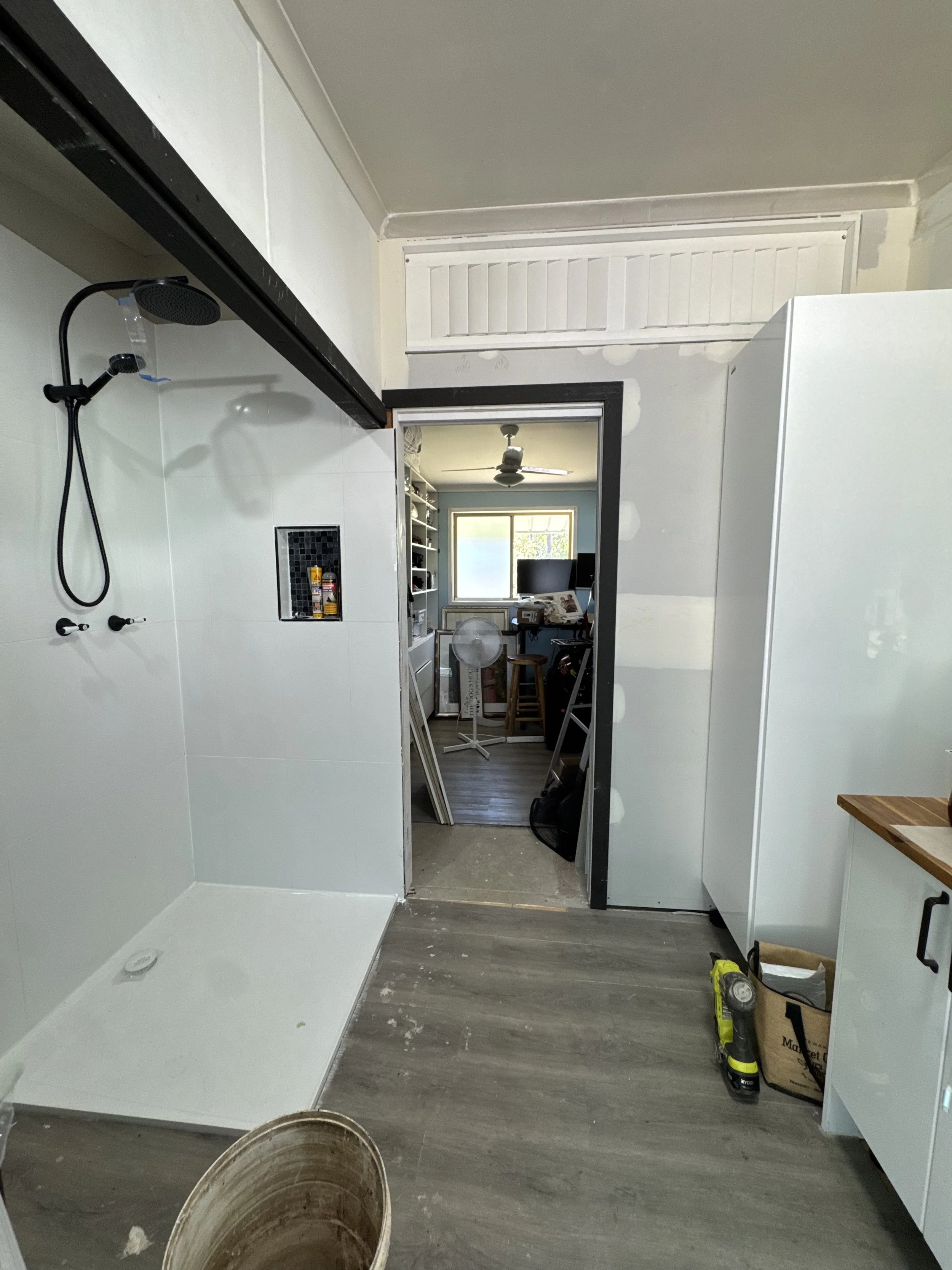 Bathroom under renovation with white tiled shower area on the left, an emerging doorway leading to a room with a closet and window, construction tools and supplies on the floor.