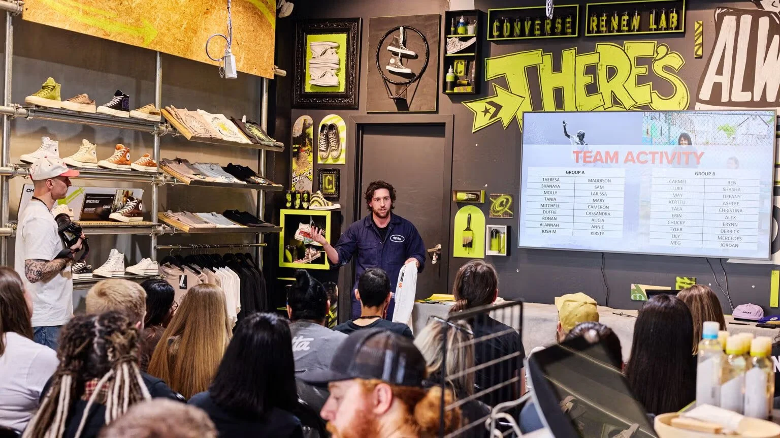 A presenter speaks to an audience in a sneaker shop or showroom, with shelves of shoes, skateboard decks, and artwork on the walls. A large screen displays a team activity list.
