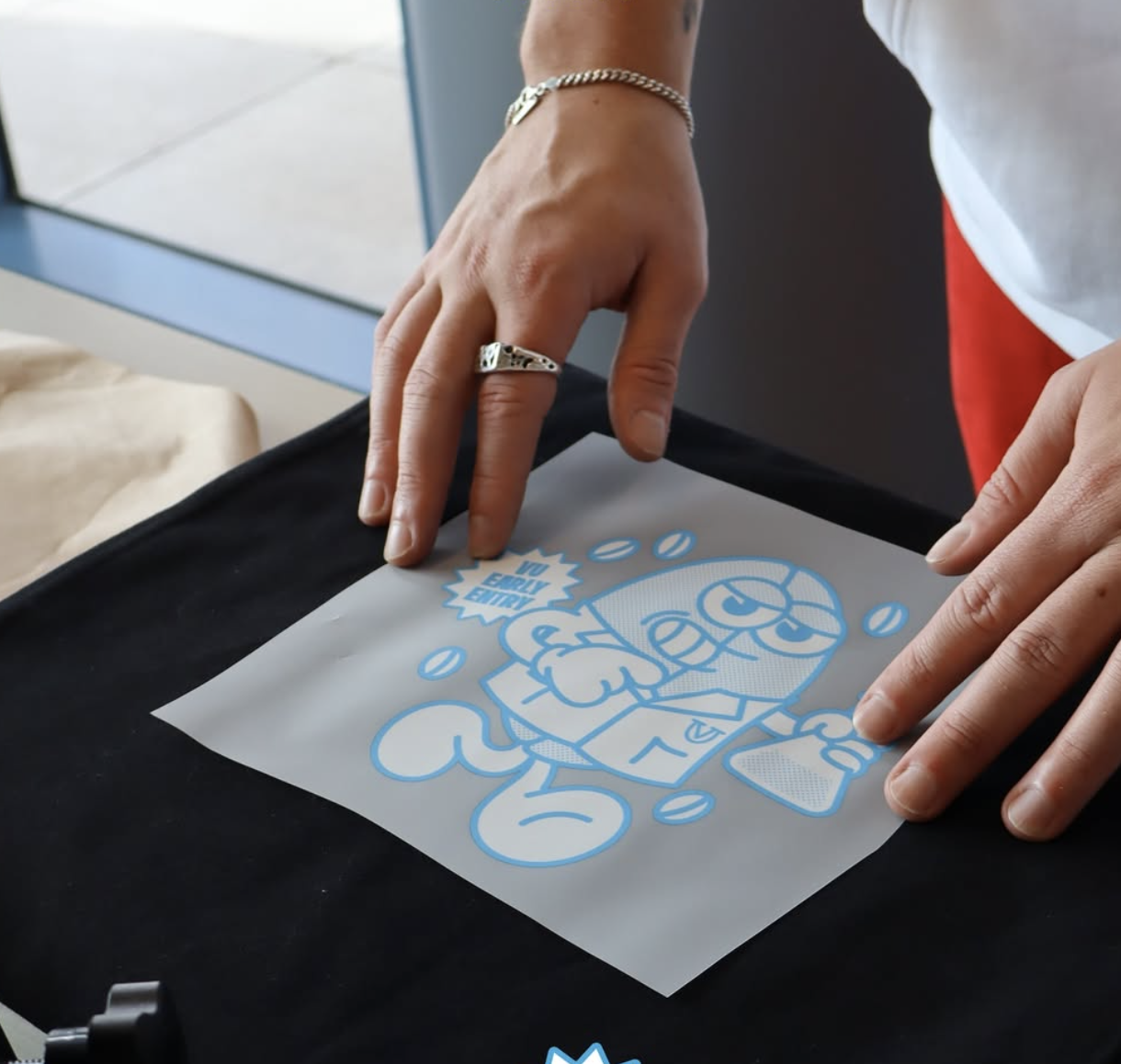 Person handling a sheet of protective film with a cartoon image of a robot and the words 'Just Eat' on it, with multiple coffee bean symbols, on a black surface.