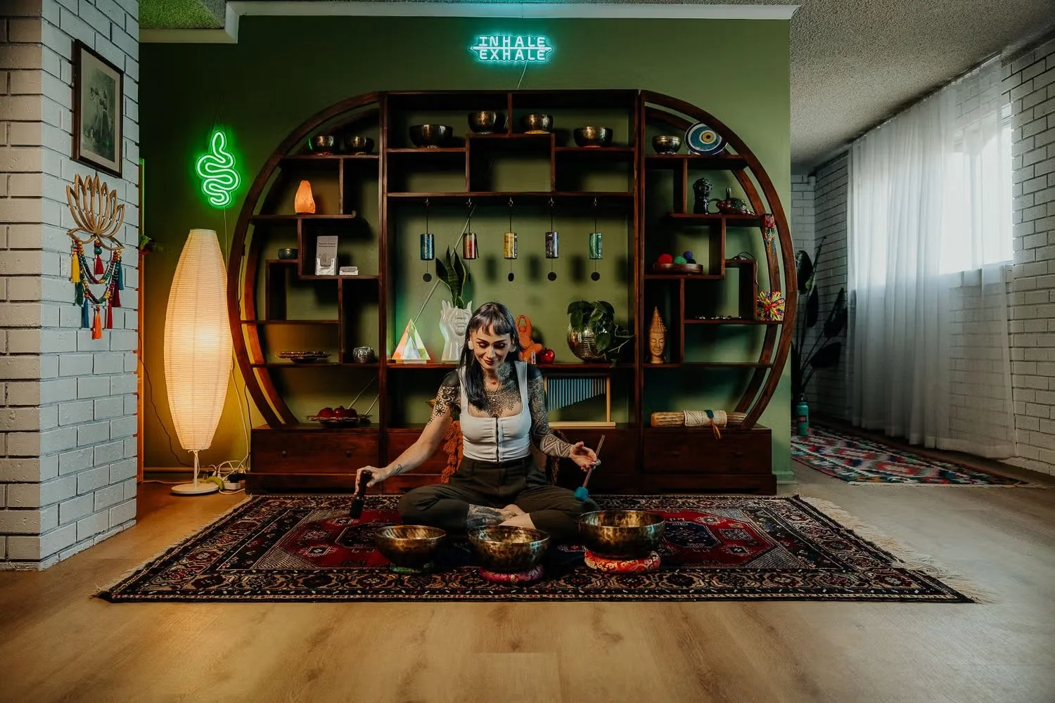 A woman sitting cross-legged on a patterned rug, playing crystal singing bowls, inside a cozy room with green walls, a large decorative wooden shelf, and neon signs that say 'INHALE EXHALE' and a snake. The room is decorated with plants, ornaments, and flowing curtains.