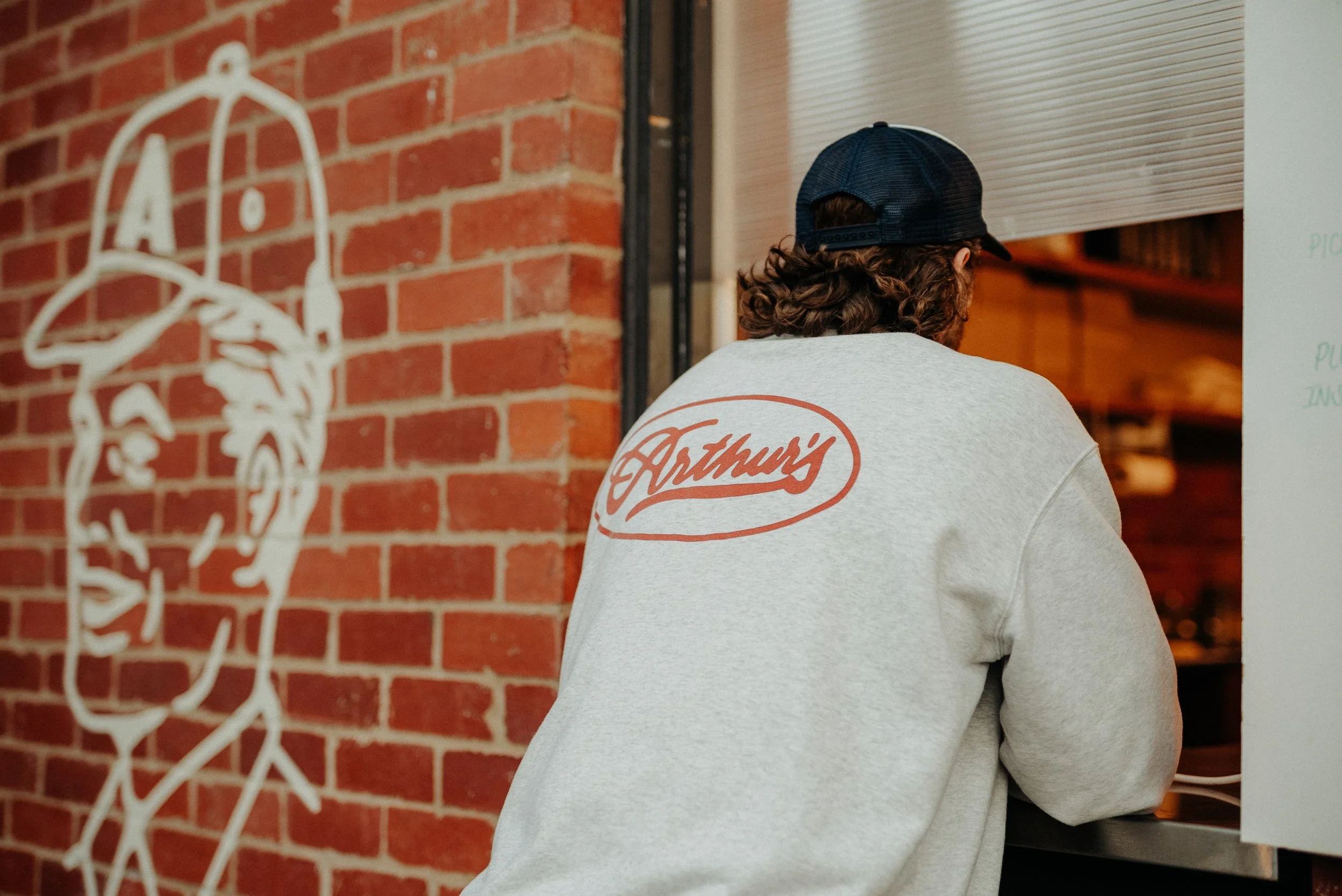 A person with curly hair wearing a gray Arthur's sweatshirt and a navy blue cap, facing away and standing near a brick wall decorated with a white outline of a baseball player