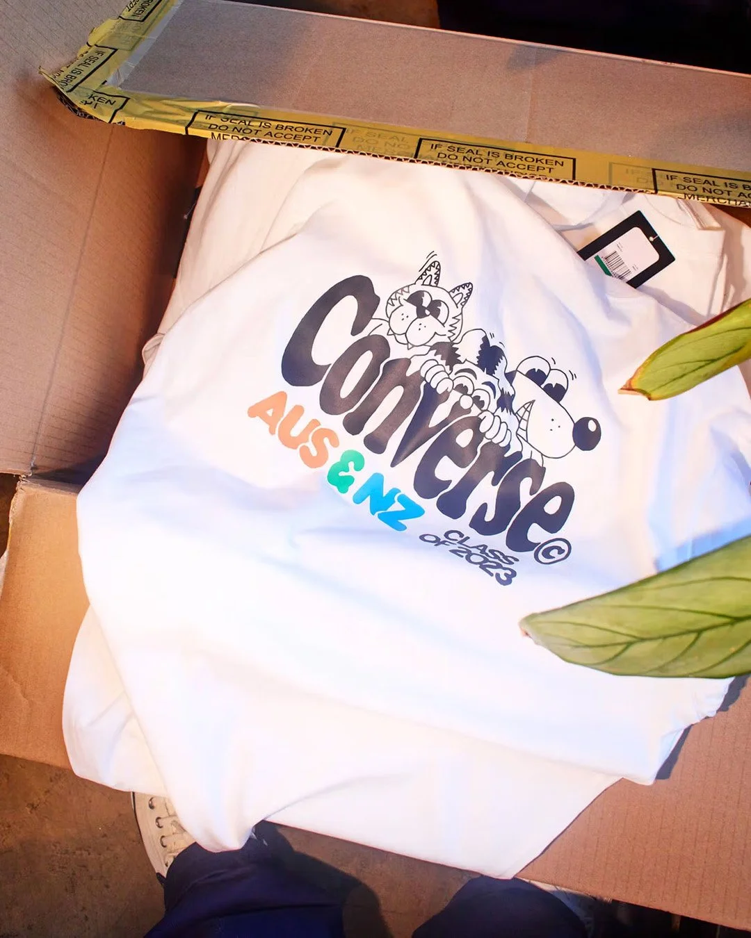 A white T-shirt with the comic strip logo for 'Comic Escape,' featuring cartoon animals and the words 'AS & NZ' and 'CLASS OF 2023,' partially inside a cardboard box.