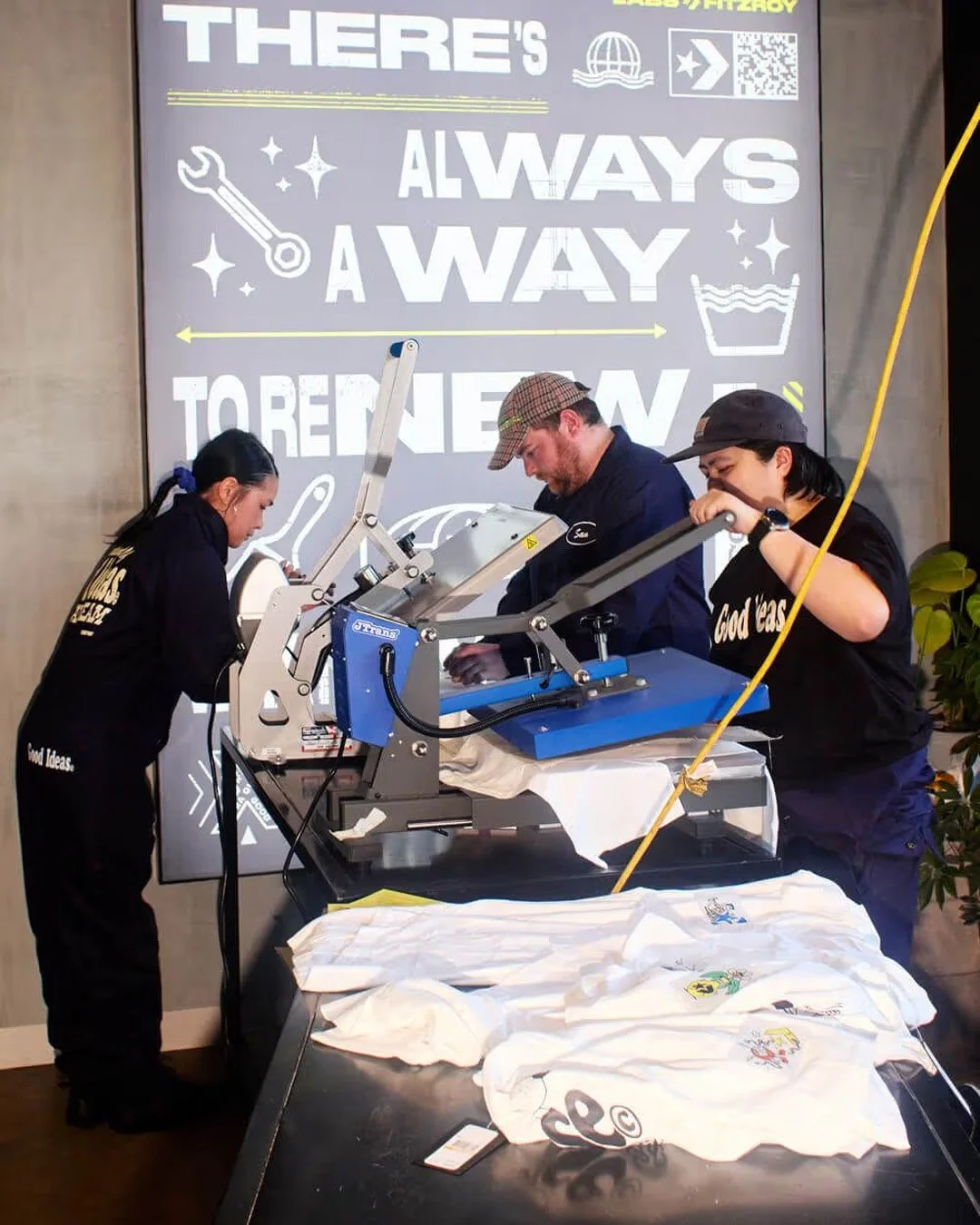 Three people working with a heat press machine, printing on white T-shirts with various logos. Behind them, a large wall sign reads 'THERE'S ALWAYS A WAY TO BE NEW' with icons of a wrench, sparkles, a globe, a plane, and a laundry basket.