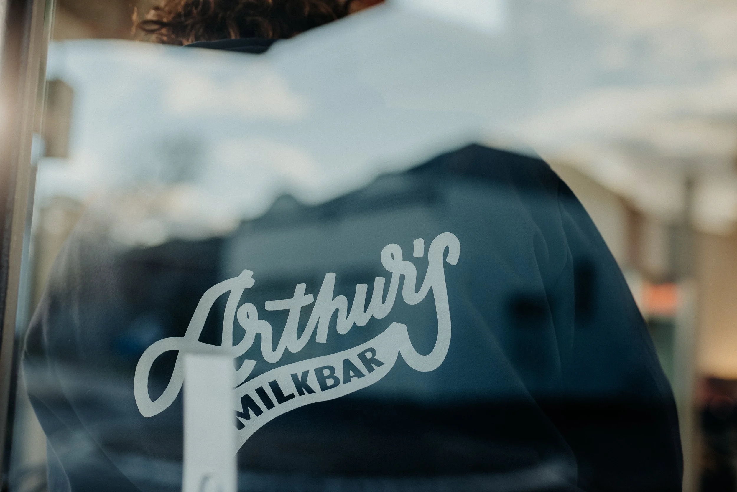 Close-up of a window sticker with the words "Breakfast, Lunch, Milkbar" in white cursive font, partially obscuring a dark jacket worn by a person with curly hair, behind the glass.