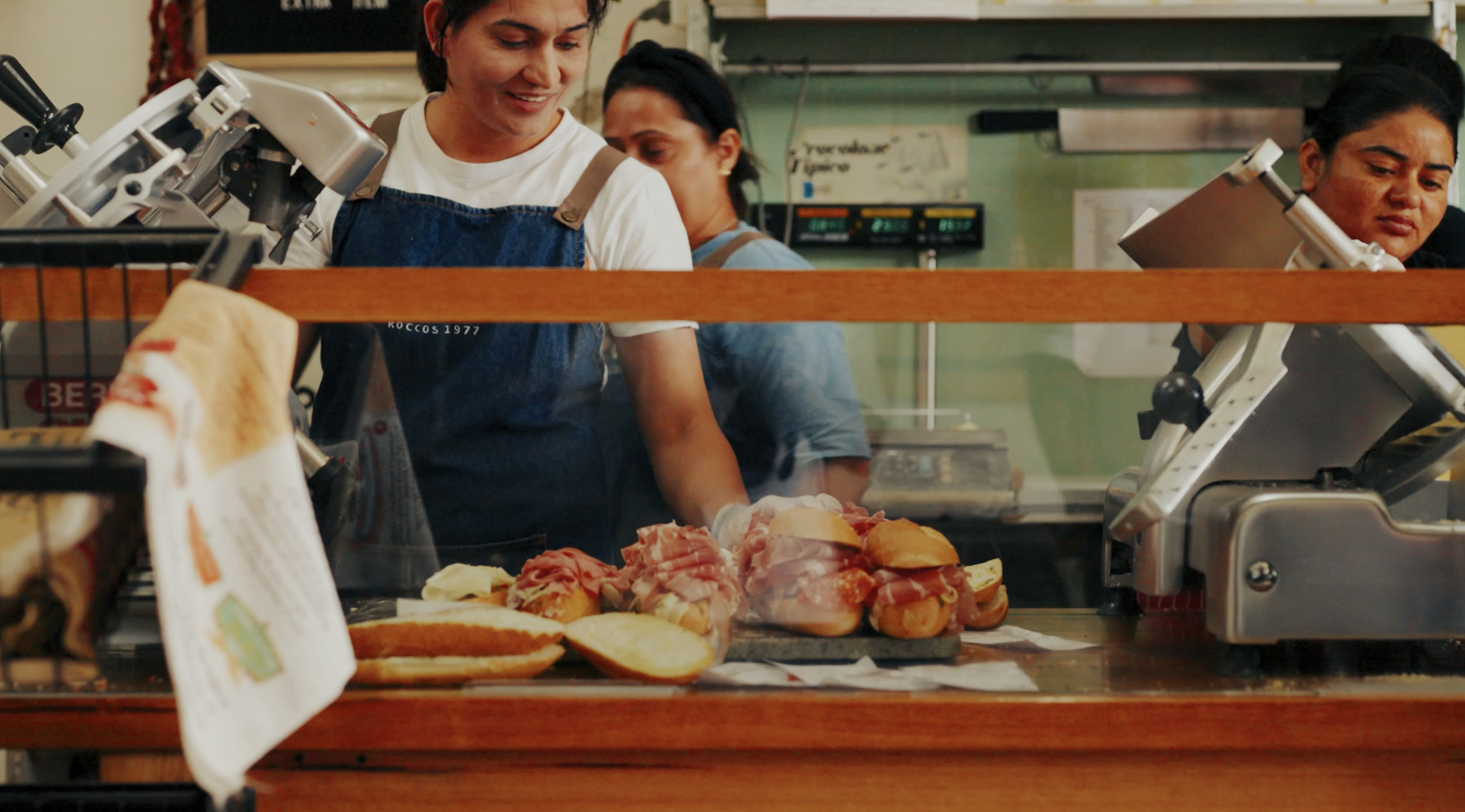 Women working at a deli counter with various meats and sandwiches on display, viewed through a glass barrier.