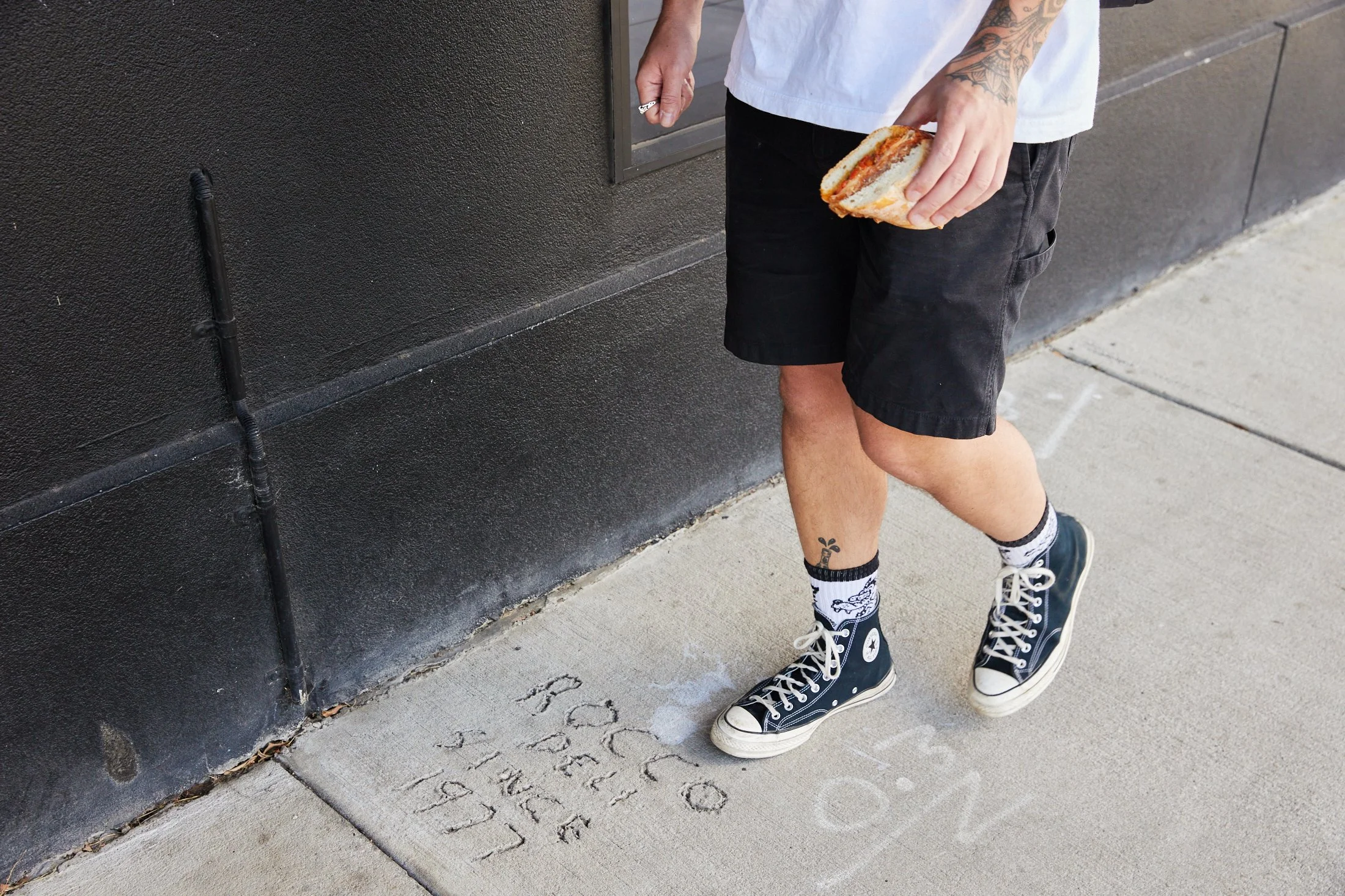 Person wearing black sneakers, socks with white cartoon characters, black shorts, white t-shirt, holding a sandwich, standing next to a black wall with writing etched into a concrete sidewalk.