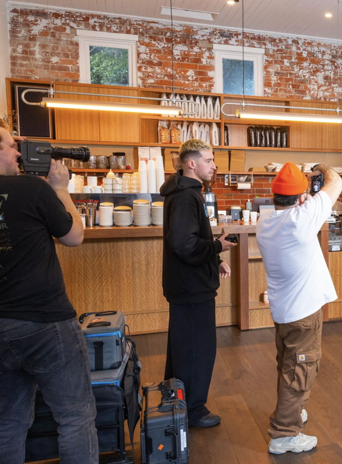 Three men are taking photos or videos inside a coffee shop with a brick wall and wooden shelves. One man in a black hoodie stands near the counter, another in a white shirt and orange beanie holds a camera, and the third man on the left holds a video