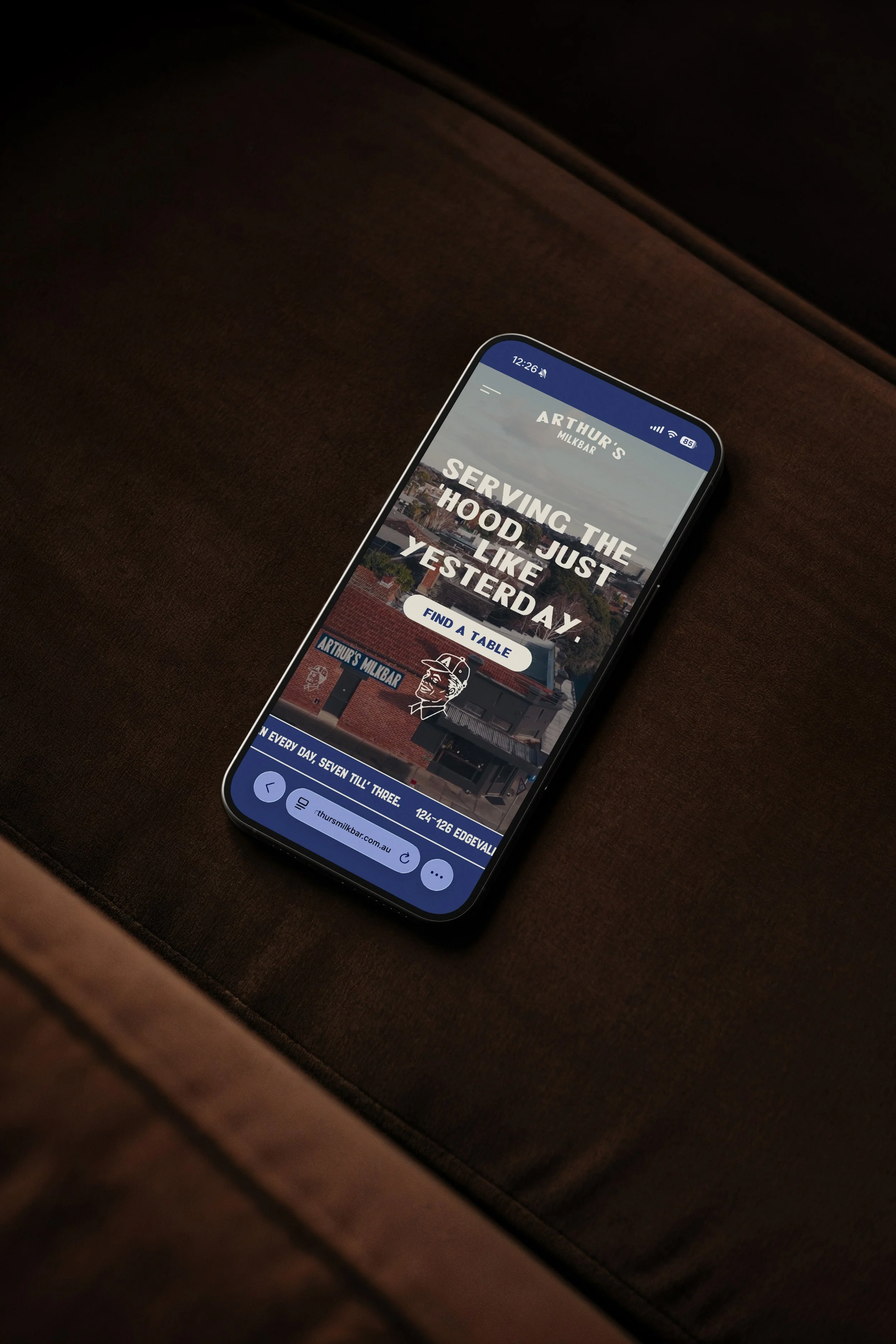 Close-up of a smartphone resting on a dark brown upholstered surface, displaying a website for Arthur's Milkbar with a message about serving the hood just like yesterday.
