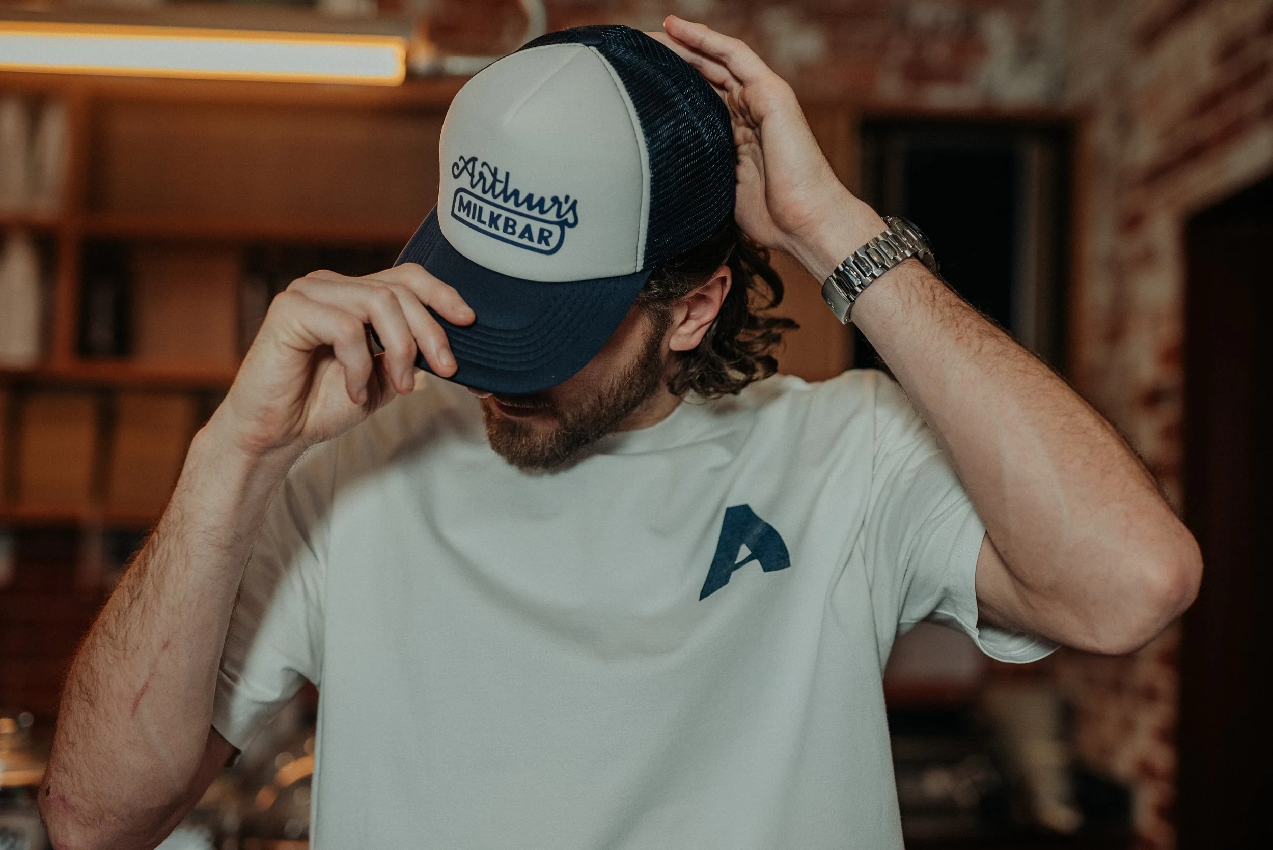 A man wearing a white t-shirt and a silver watch adjusting a trucker hat with a logo that reads 'Arthur's Milkbar' in an interior space with brick walls and wooden shelves.
