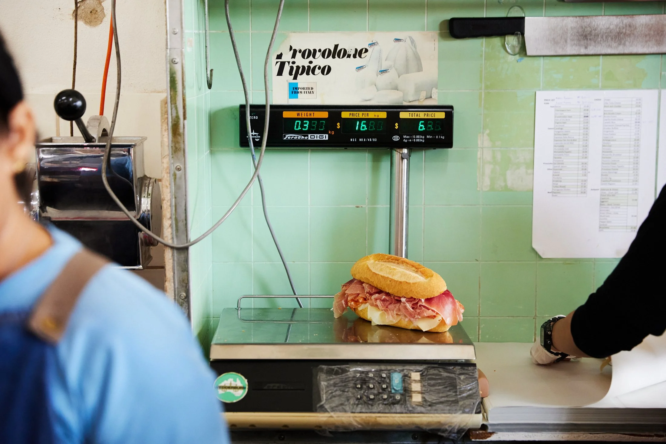A sandwich with ham and cheese on a baguette bun placed on a green counter in front of a scale displaying weight, price per kilogram, and total price, in a deli or sandwich shop with green tiled walls.