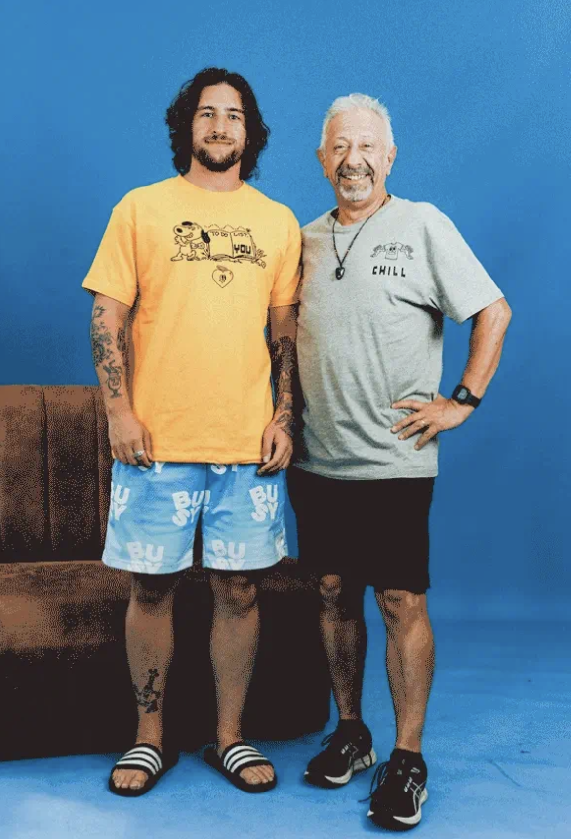Two men standing indoors against a blue background. The man on the left has long dark hair, a beard, and tattoos on his arms. He's wearing a yellow T-shirt with a cartoon dog and the words "TO DO" and "YOU" on it, blue shorts with white text, and bla