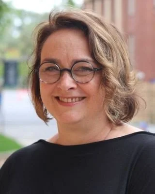 Portrait of a smiling woman with wavy brown hair and round glasses wearing a black top outdoors