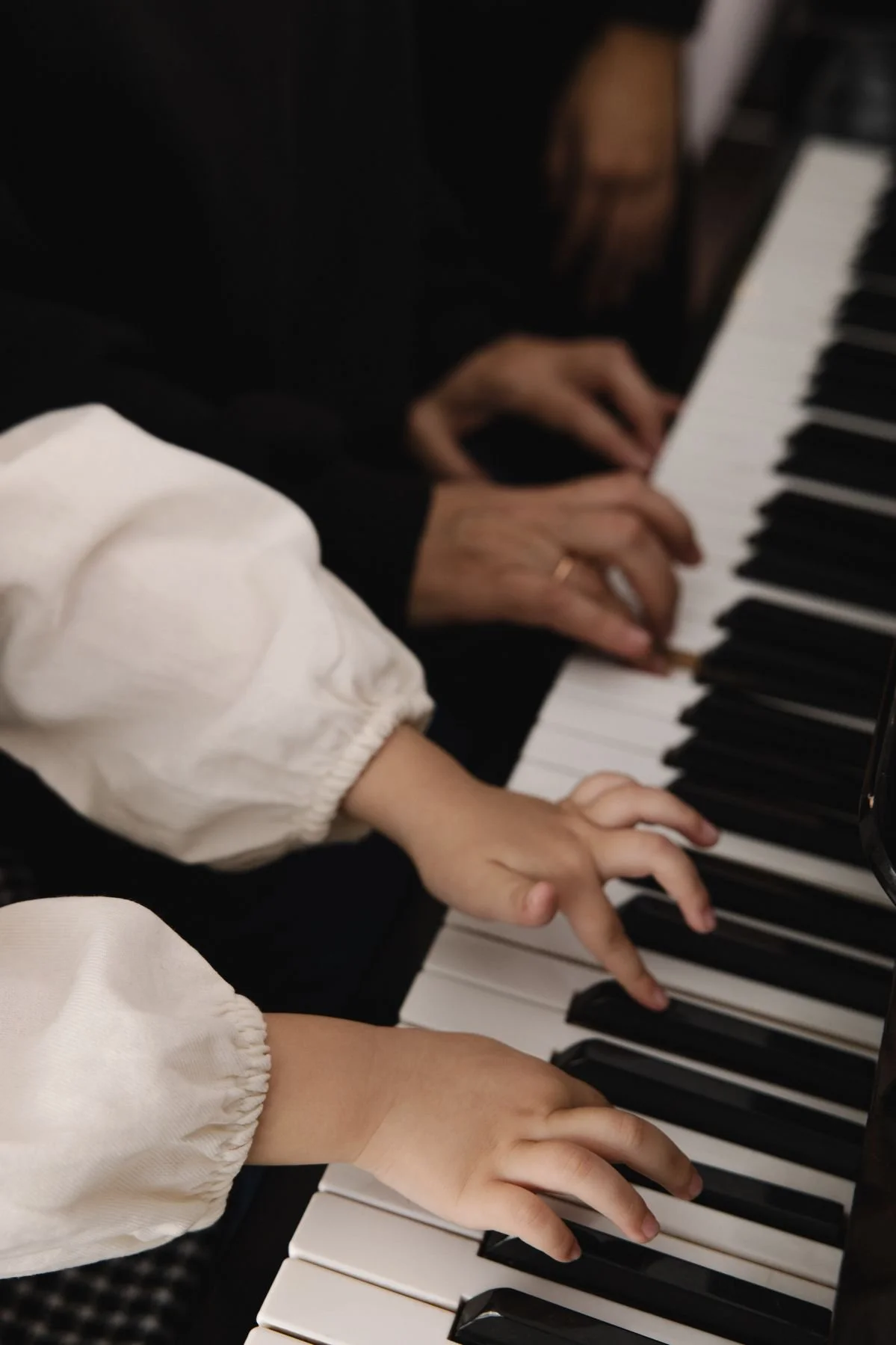 Close-up of two children and an adult playing the piano together. The children are wearing white long-sleeved shirts, and the adult is wearing a black shirt. The focus is on their hands on the keys of a black and white piano.