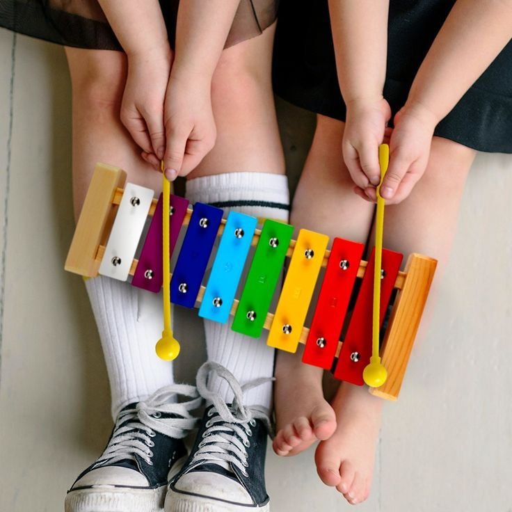 Two children sitting with their legs extended, playing a colorful xylophone. The children are wearing school uniforms and socks, and the xylophone is resting on their laps.