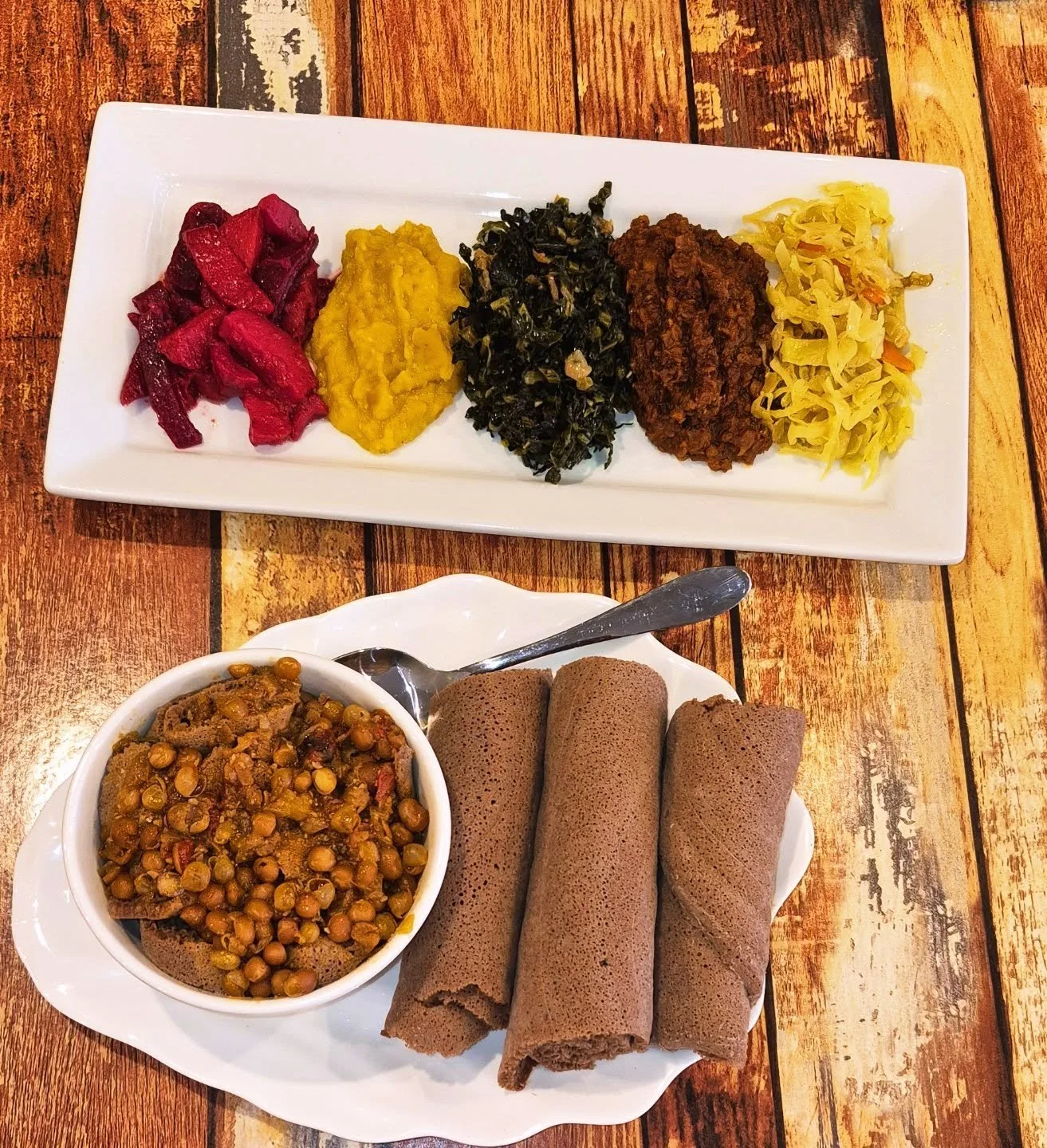 A plate with various Ethiopian foods including lentil stew, rolled injera, a bowl of chickpeas and vegetables, and a side of mixed vegetables on a wooden table.