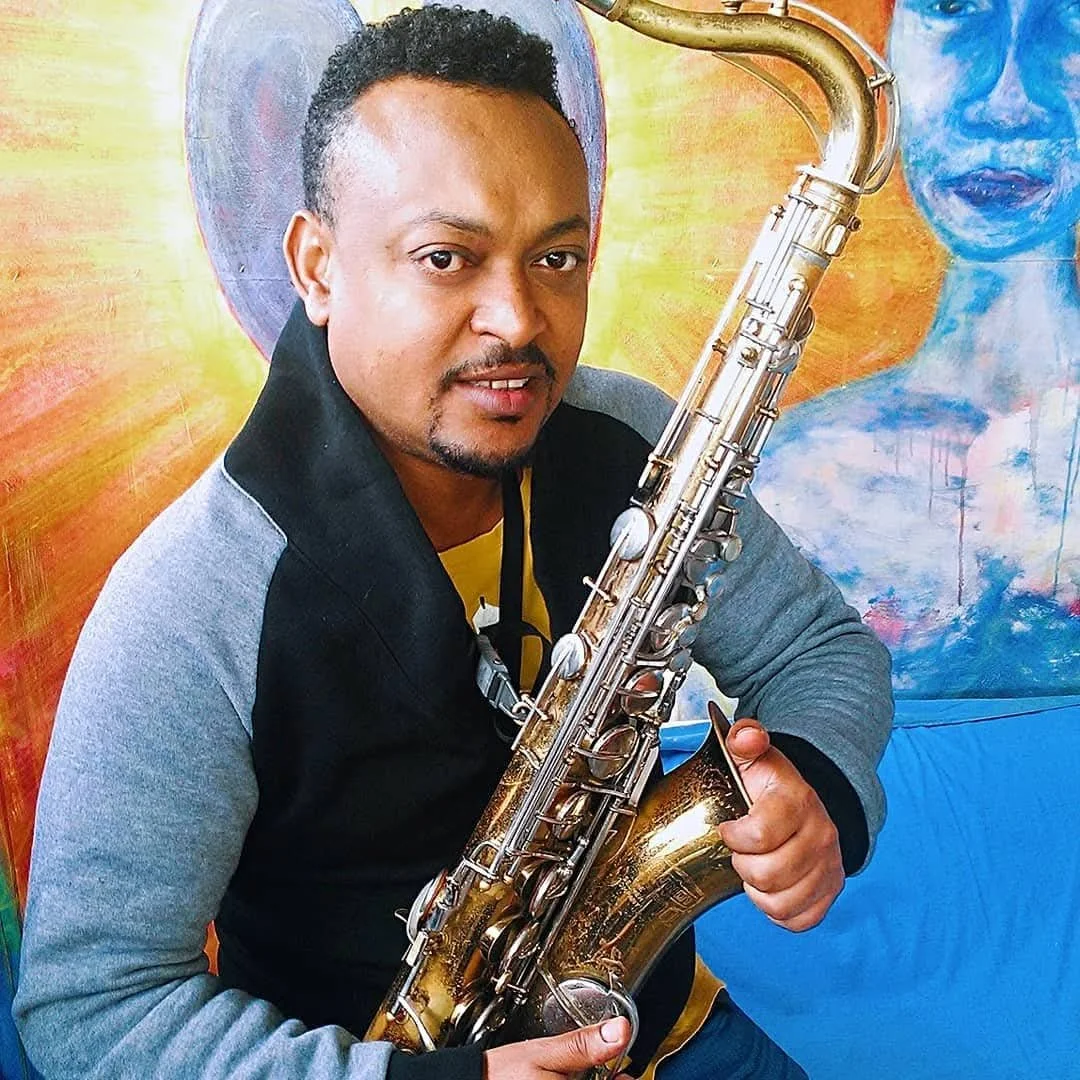 A man holding a saxophone, sitting in front of colorful art with a woman's face in shades of blue and orange.