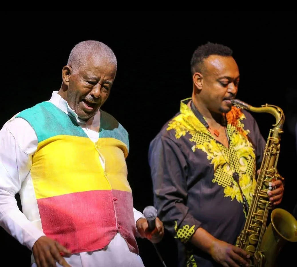 Two men performing on stage: one singing and holding a microphone, the other playing a saxophone. The singer wears a colorful shirt with green, yellow, and red stripes; the saxophonist wears a black shirt with yellow embroidery.