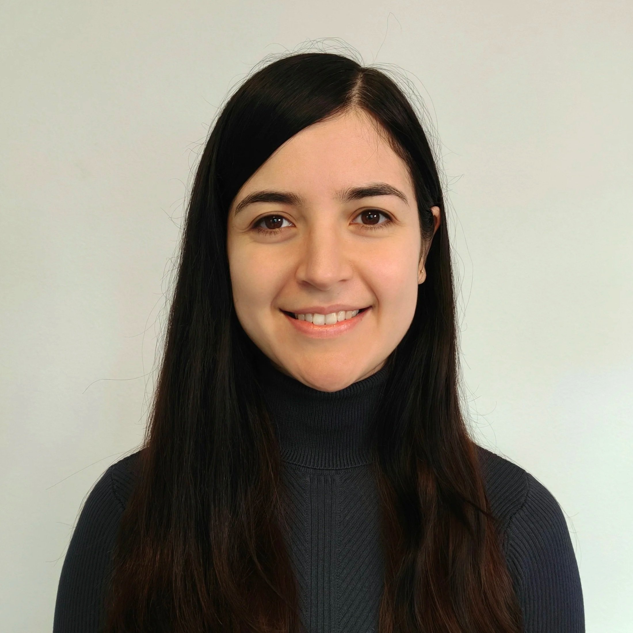 A woman with long dark hair, brown eyes, and light skin, smiling, wearing a black turtleneck, against a plain light gray background.