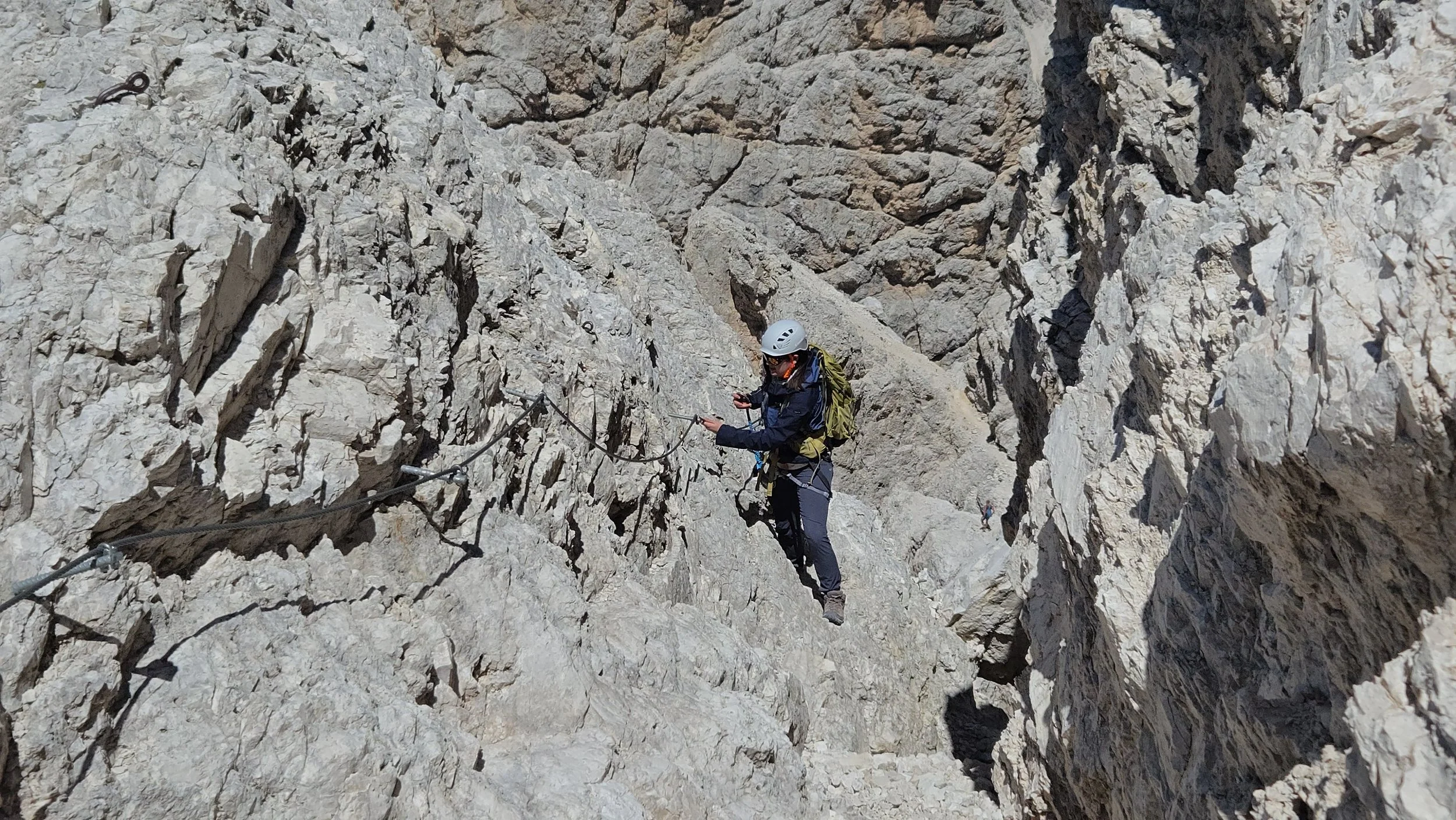 A person wearing a helmet and harness descending a steep rocky mountain trail with via ferrata safety cables.