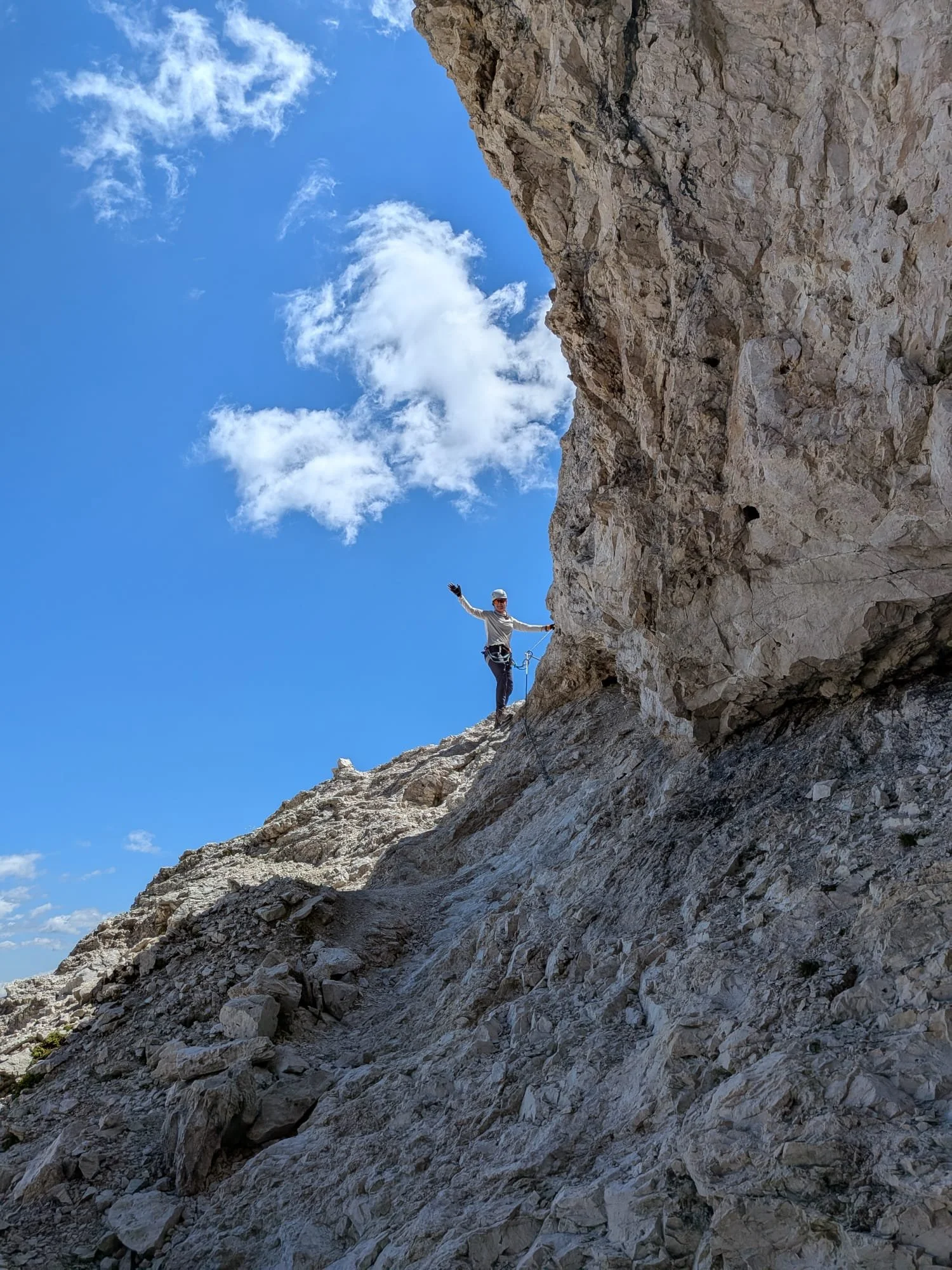 A person wearing a helmet and climbing gear standing on a rocky mountain slope near a tall rock face, with a blue sky and some clouds in the background.