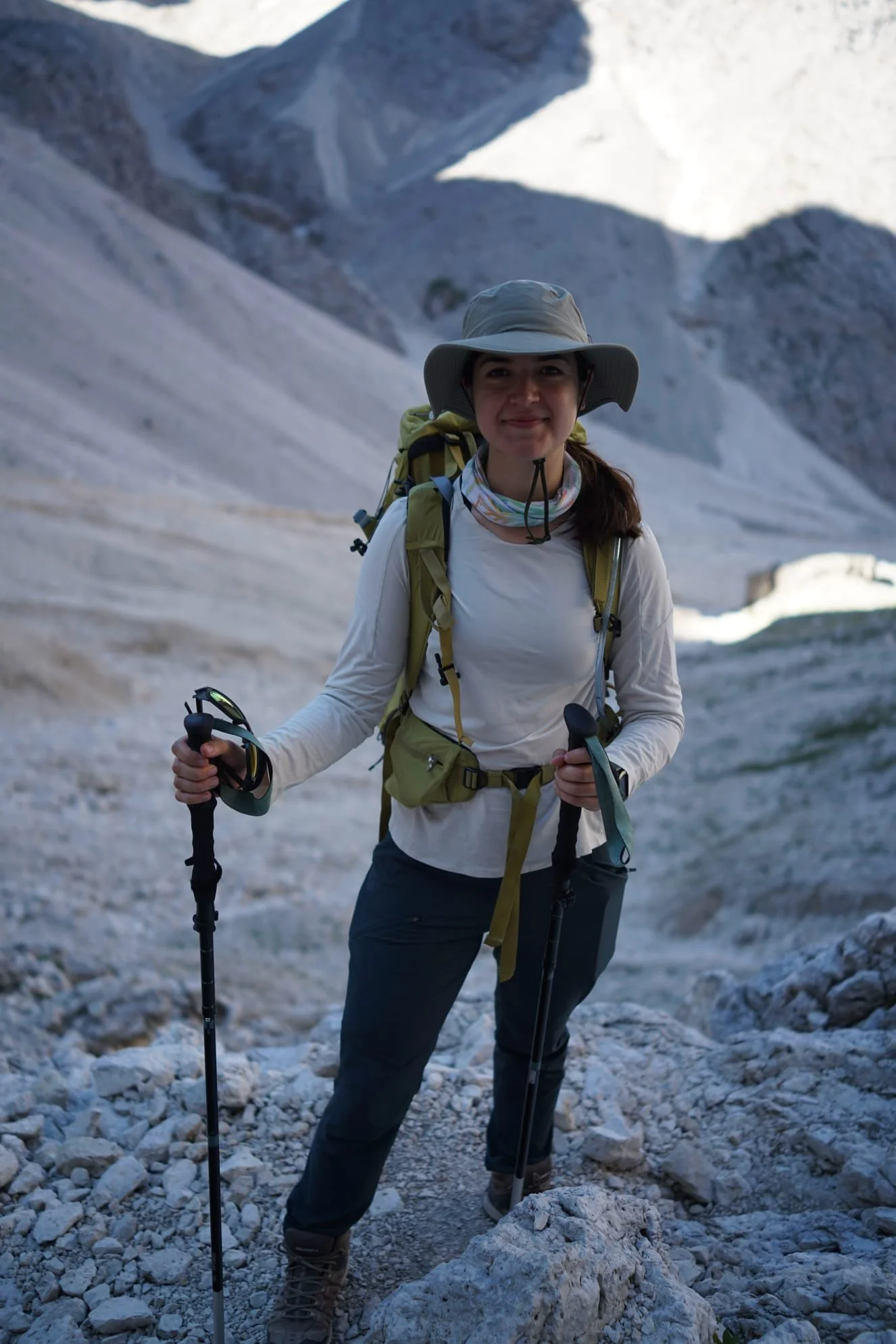 Woman hiking in rocky mountainous terrain with a backpack, hiking poles, and wearing a wide-brimmed hat, in a remote outdoor setting.