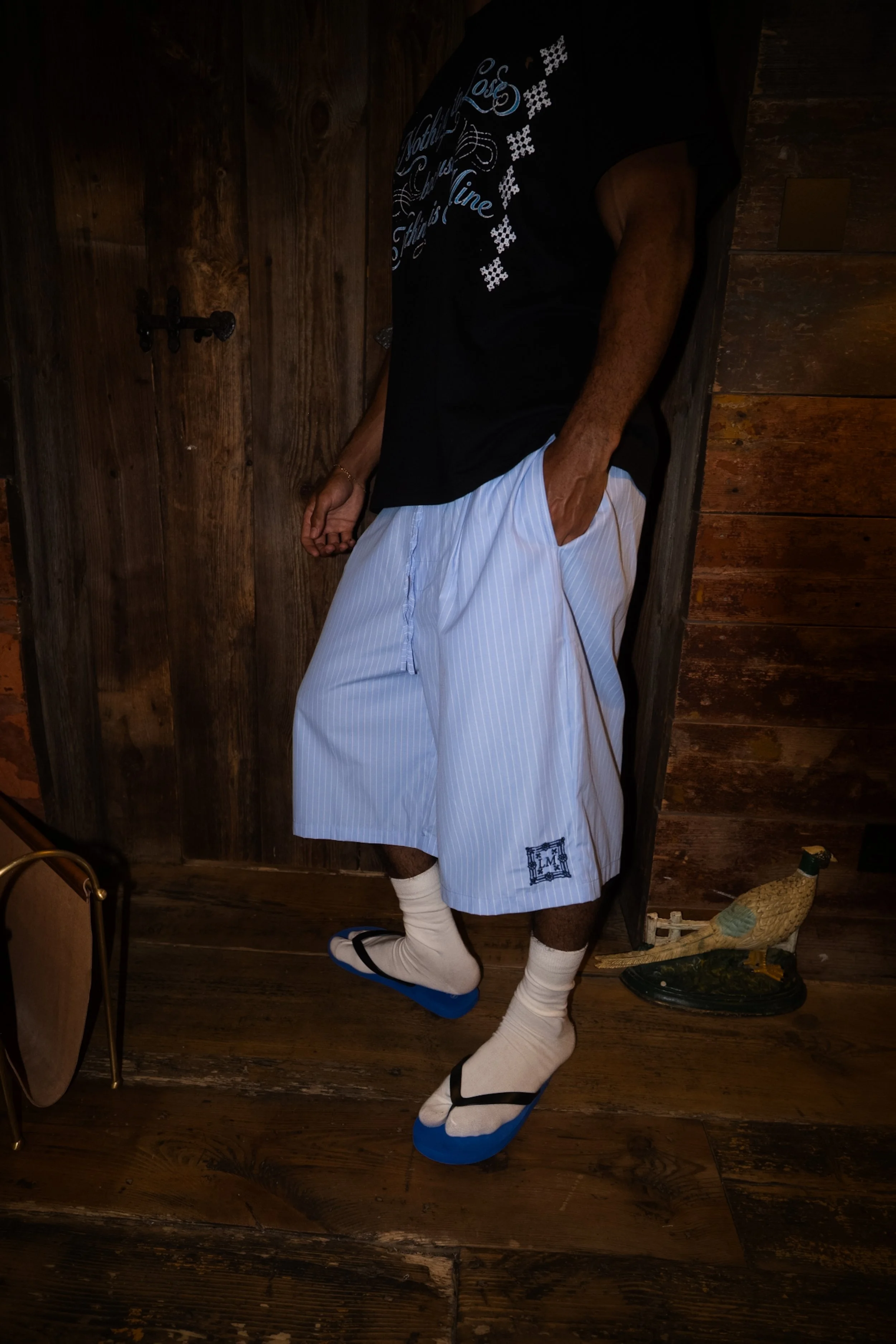 A person standing in front of a wooden wall, wearing a black T-shirt, white and blue striped shorts, white socks, and traditional Japanese wooden sandals called geta.