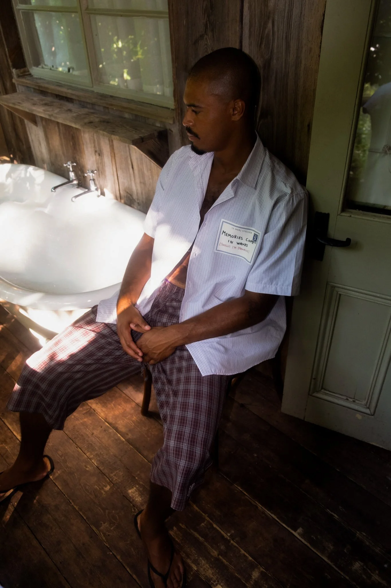 A man wearing a striped shirt and plaid shorts sitting in front of a sink with a window behind him, on a wooden floor with dark wood planks.