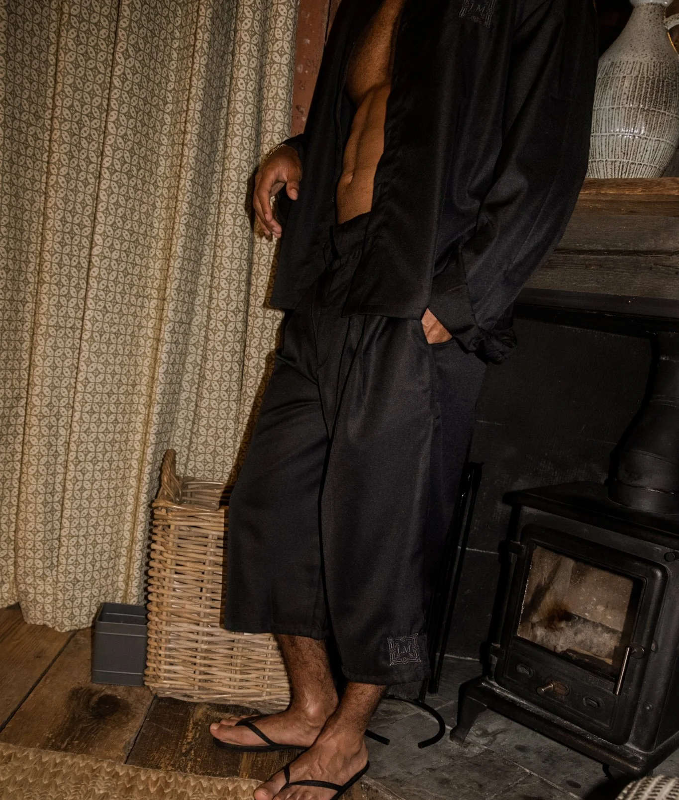 A person wearing a black satin shirt open at the front, black shorts, and flip-flops, standing indoors near a wicker basket and a fireplace with a wooden mantle.