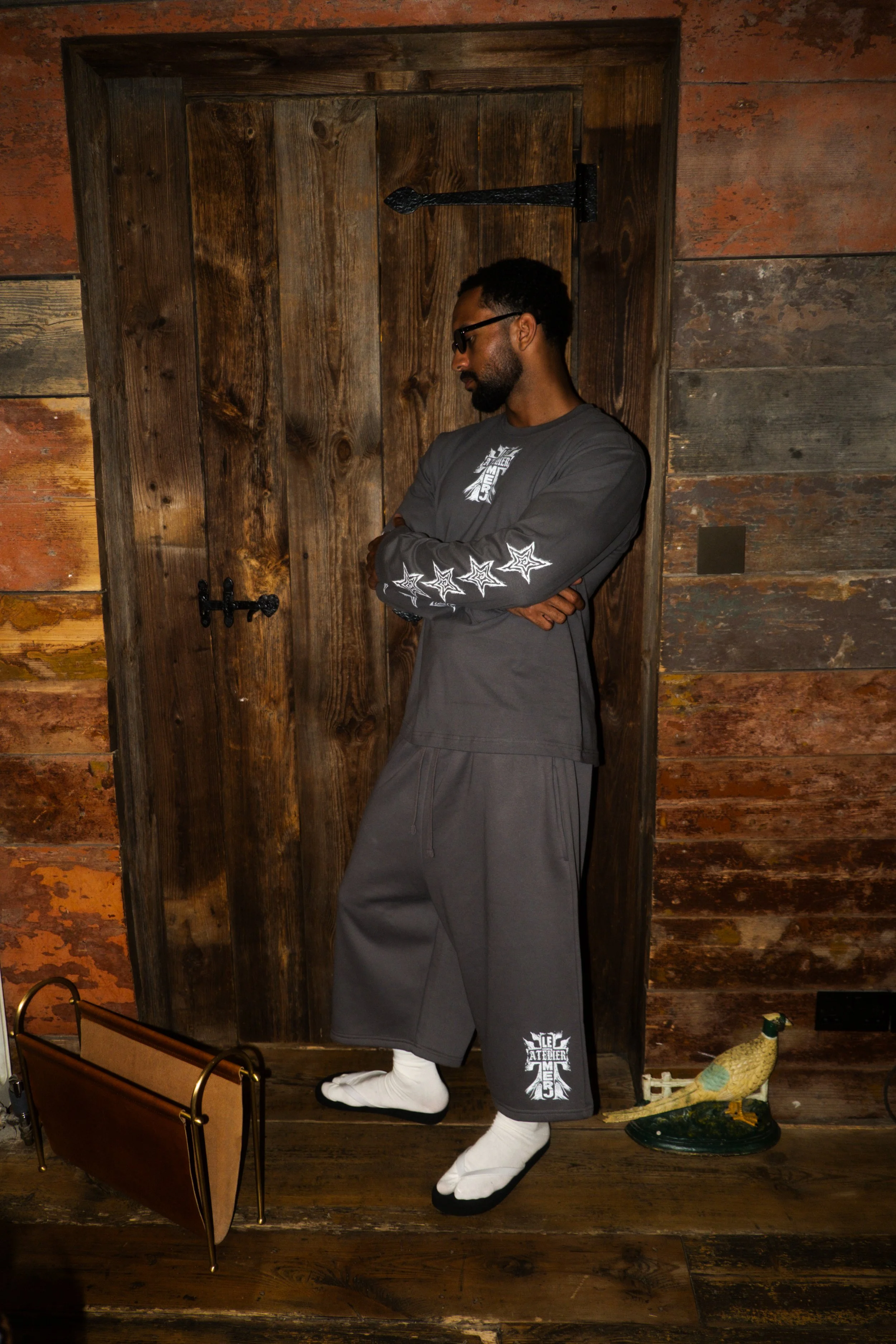 Man wearing gray long-sleeve shirt with star designs on the sleeves and loafers, standing with arms crossed against a wooden wall with a closet door behind him.