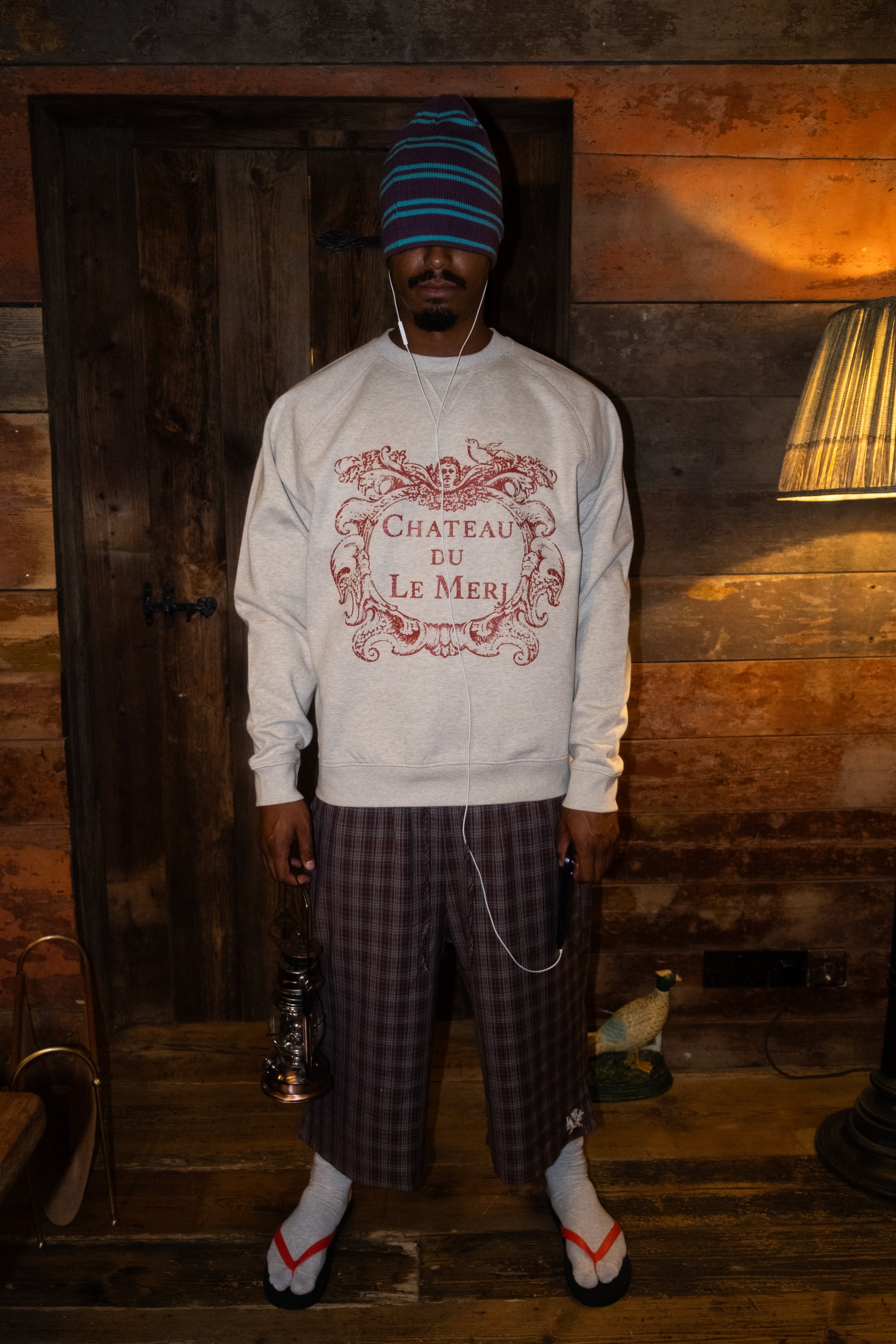 A man with a hidden face wearing a beanie and wearing a sweatshirt with the text 'Chateau du Le Meri' standing indoors in front of a wooden wall, holding a smoking hookah, with socks that have white, grey, and red colors, and checkered pajama pants.