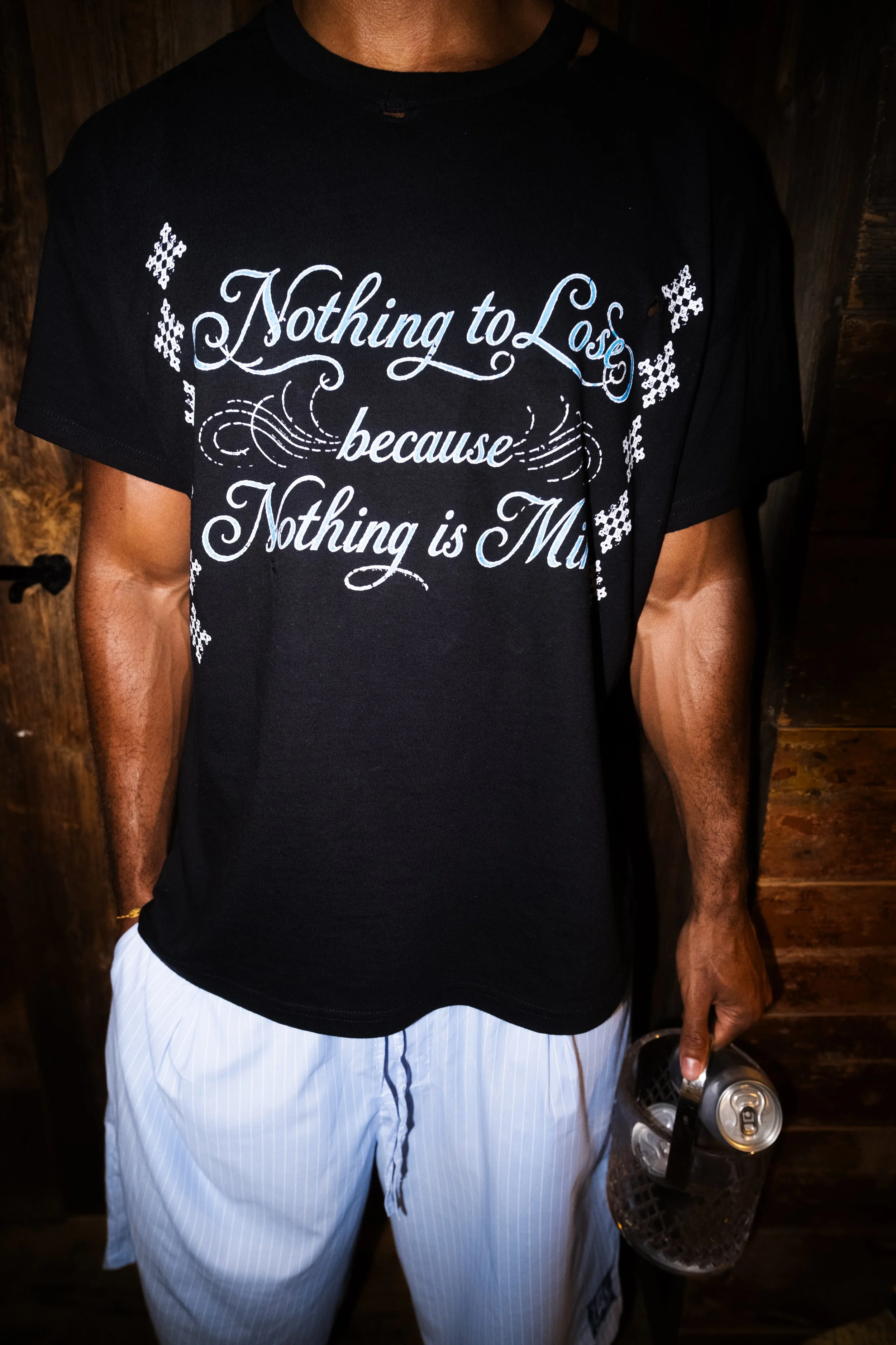 A man wearing a black t-shirt with white cursive text that says "Nothing to Lose because Nothing is Mine" and white striped shorts, holding a few cans and a black basket.