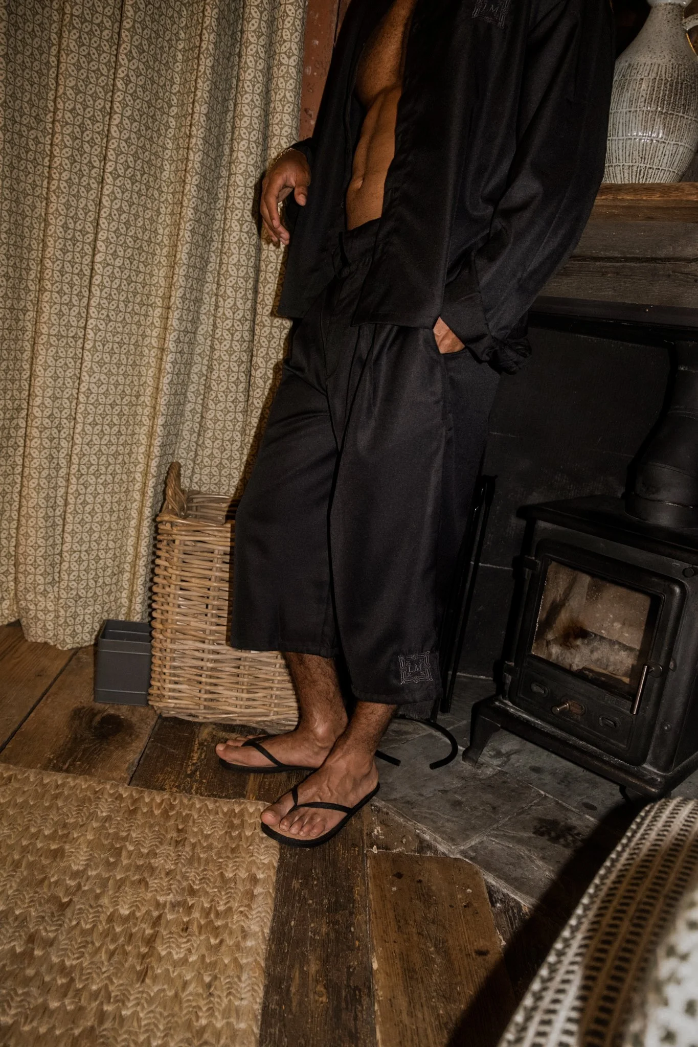 A man wearing black shorts, flip-flops, and an open black satin shirt standing indoors next to a chimney and a curtain with a floral pattern, with his hand resting on his hip.