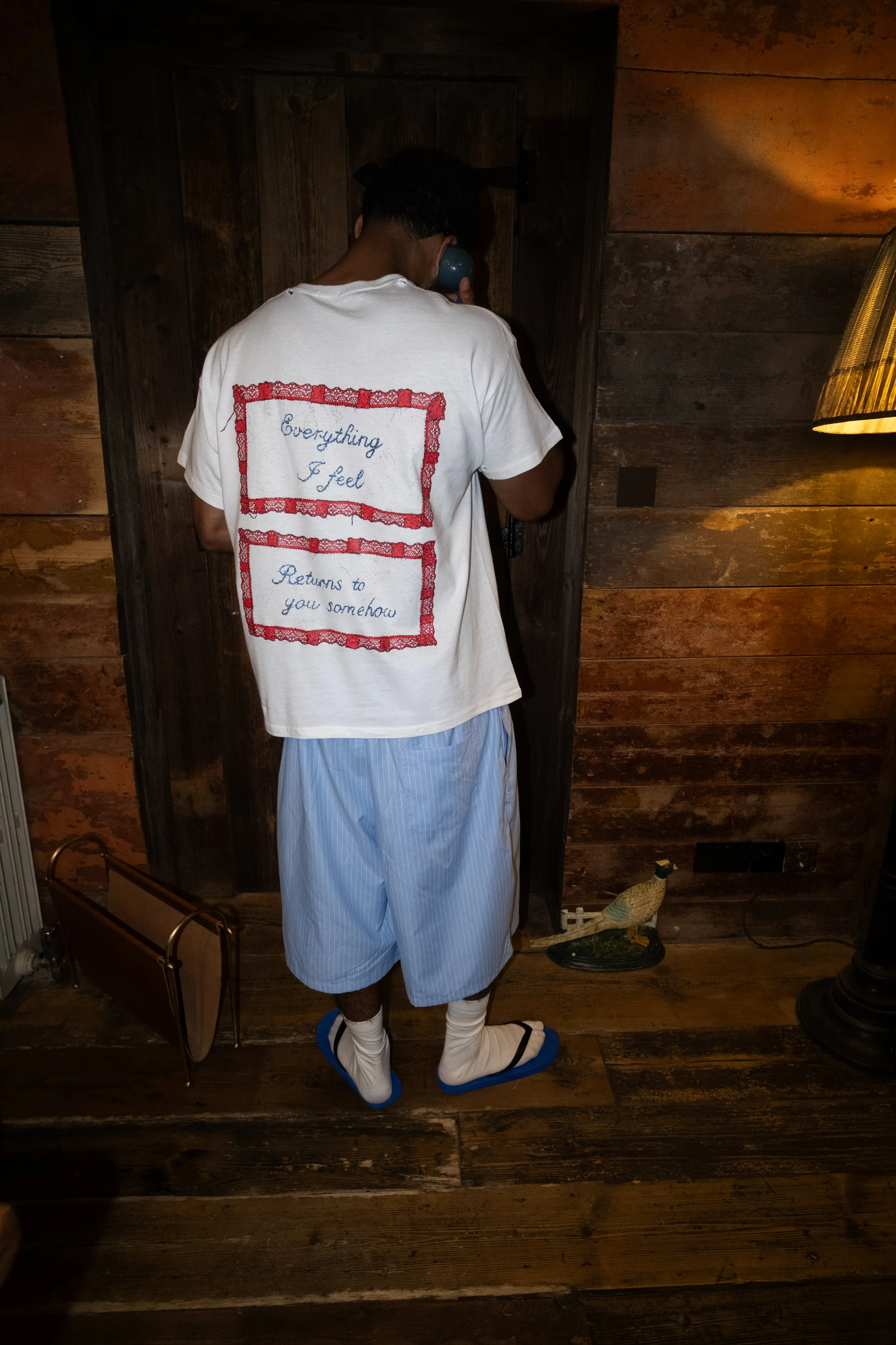 A person standing indoors on a wooden floor, wearing a white T-shirt with embroidered text and a pair of striped pajama pants. The person is holding a phone to their ear and is wearing blue slippers over socks. The room has wooden walls, a lamp on th