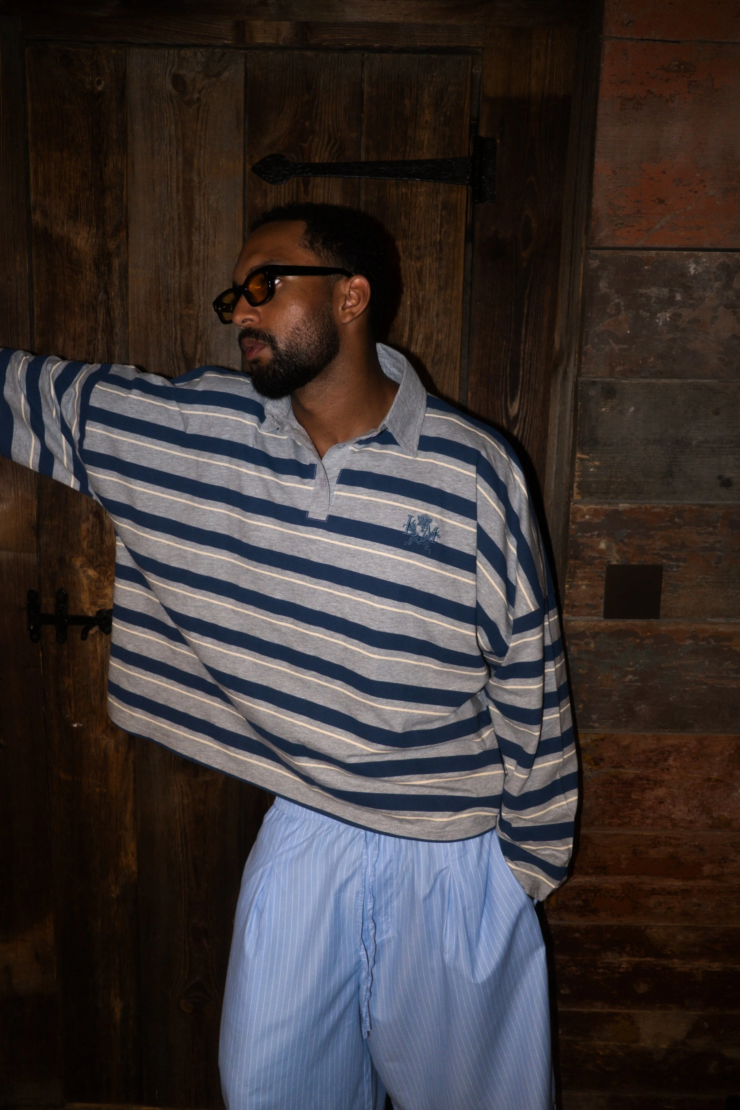 A man with a beard and dark sunglasses standing against a wooden wall, wearing a blue and white striped long-sleeve shirt and light blue striped pants.