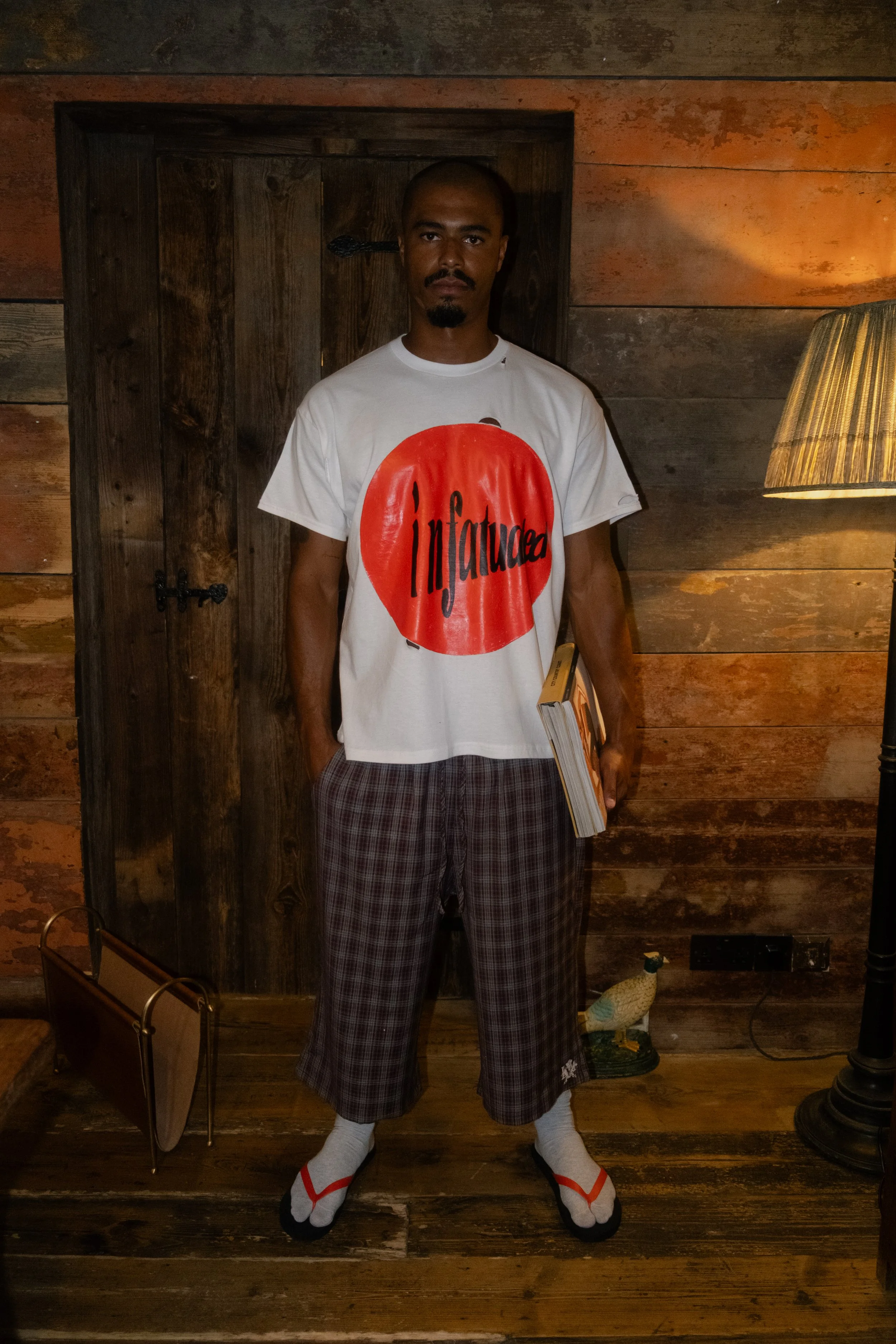 A man standing indoors in front of wooden wall, wearing a white T-shirt with a red circle and black text, plaid pants, white socks, and slippers, holding books in his left hand.
