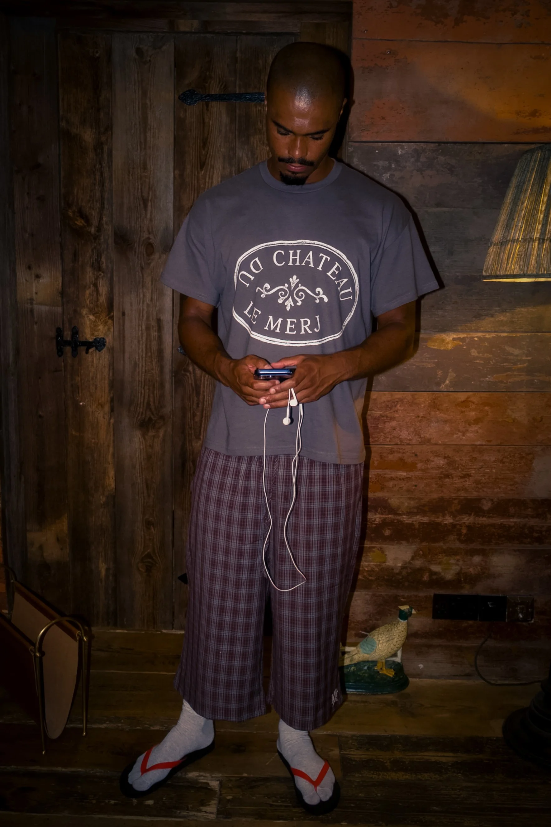 A man with a shaved head and beard stands indoors against a wood-paneled wall, looking down at his phone. He wears a gray T-shirt with white text and a design, plaid pajama pants, and is wearing socks.