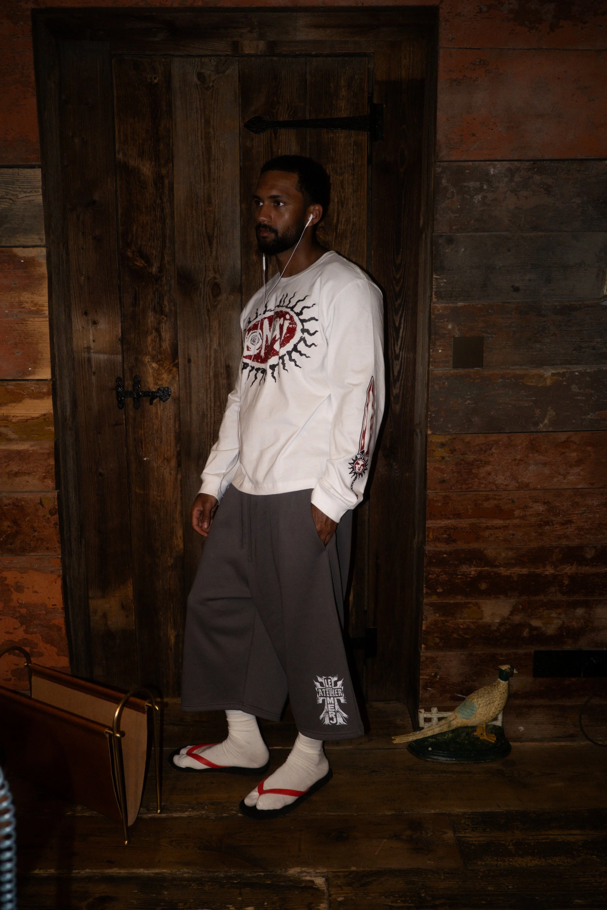 A man with dark hair and beard standing indoors against a wooden wall, wearing white earphones, a white long-sleeve shirt with a red and black graphic, dark gray shorts with white and red tabi socks, and black sandals with red straps. There is a wood