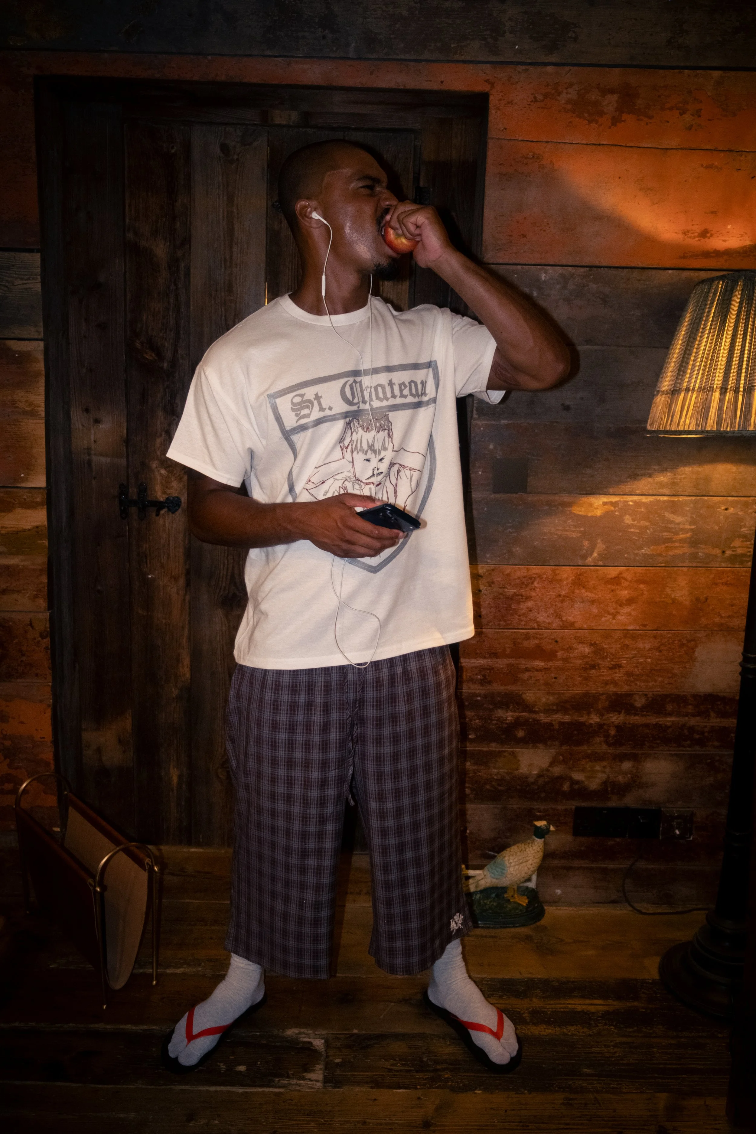 A man with a shaved head and dark skin, wearing a graphic T-shirt, plaid pajama pants, white socks, and traditional Japanese footwear, is biting into a peach. He is in a room with wooden walls and a lamp on the right side, with various decorative ite
