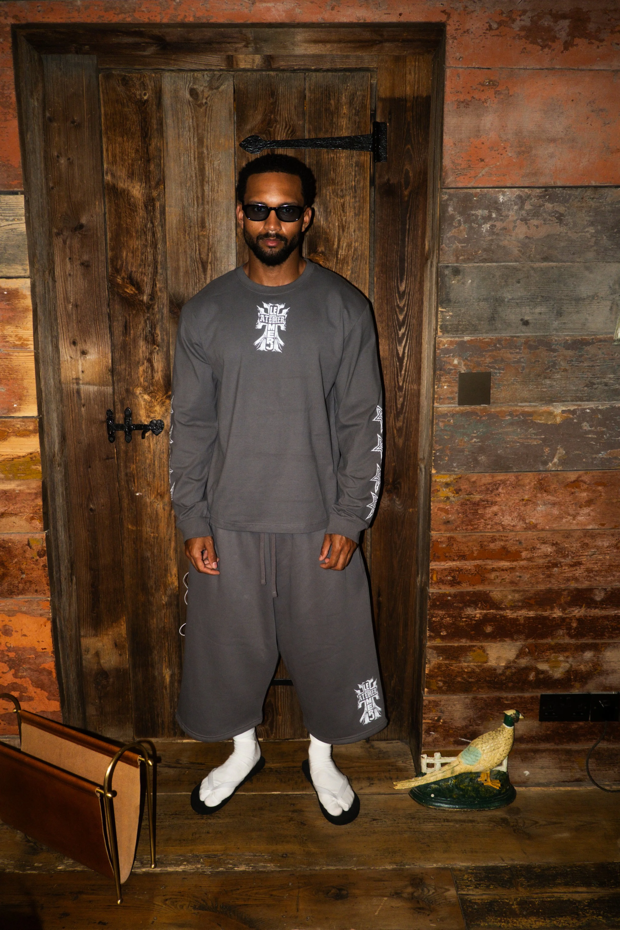 A man with dark skin, sunglasses, and a beard stands in front of a wooden wall with a door, wearing a gray long-sleeve shirt and oversized gray shorts. There is a decorative bird sculpture on the floor to his right and a brown bag with gold accents i