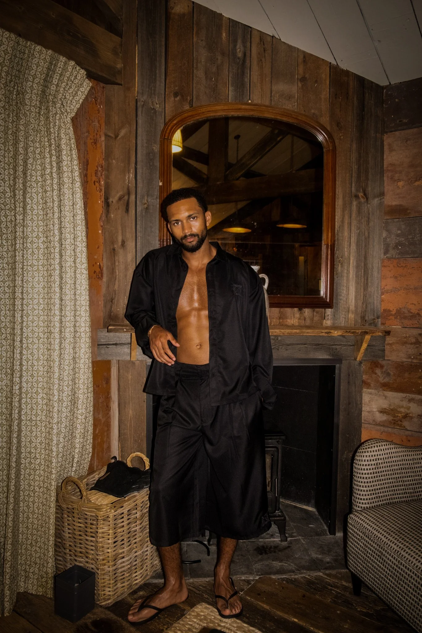 A young man with a beard and dark hair stands indoors in front of a wooden wall with a mirror. He is shirtless, wearing an open black jacket and black shorts, and sandals. The room has wood paneling, a patterned curtain, a wicker basket, a chair, and
