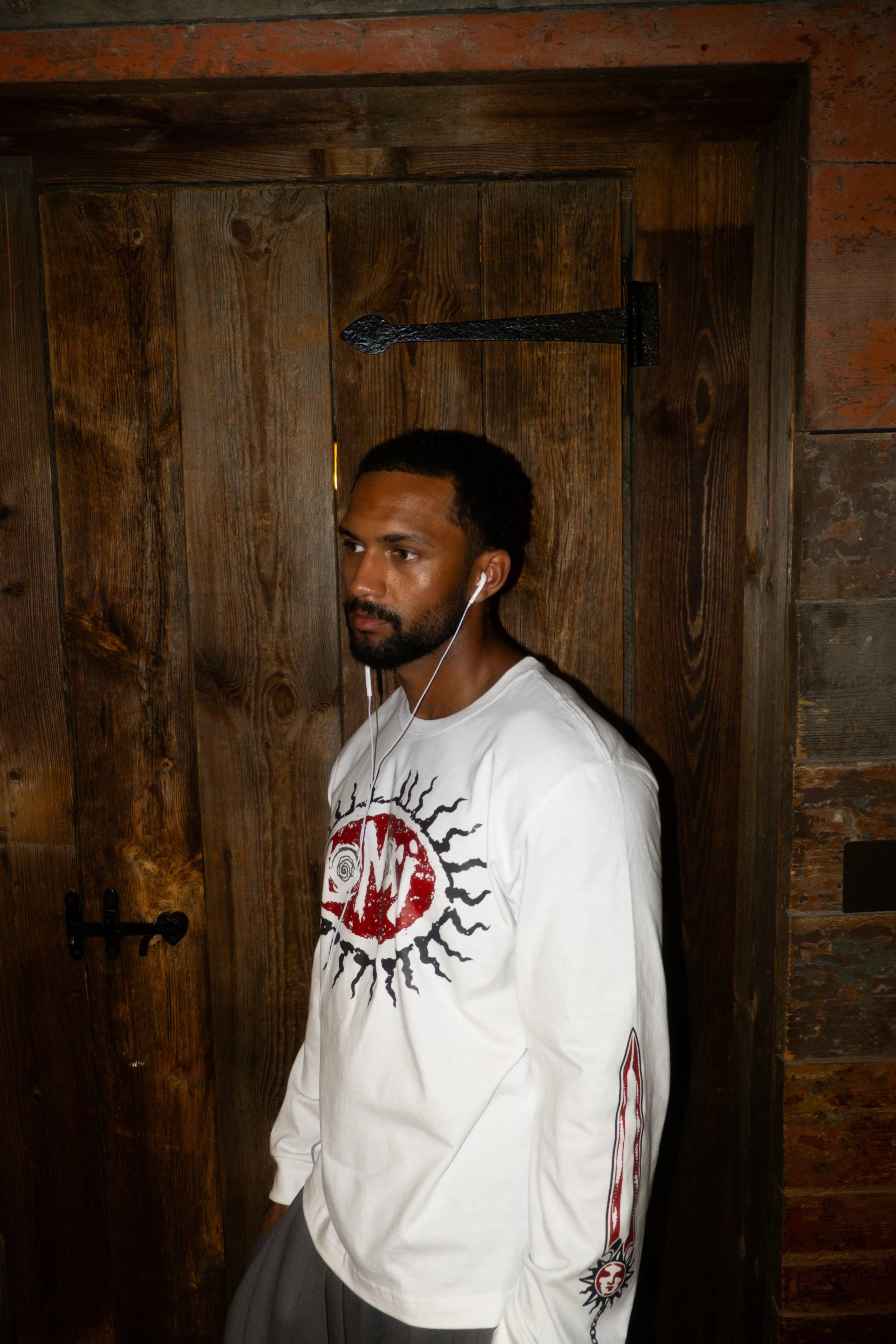 A man with dark hair, a beard, and wearing a white graphic long-sleeve shirt stands in front of a wooden door with black iron hardware in a rustic setting.
