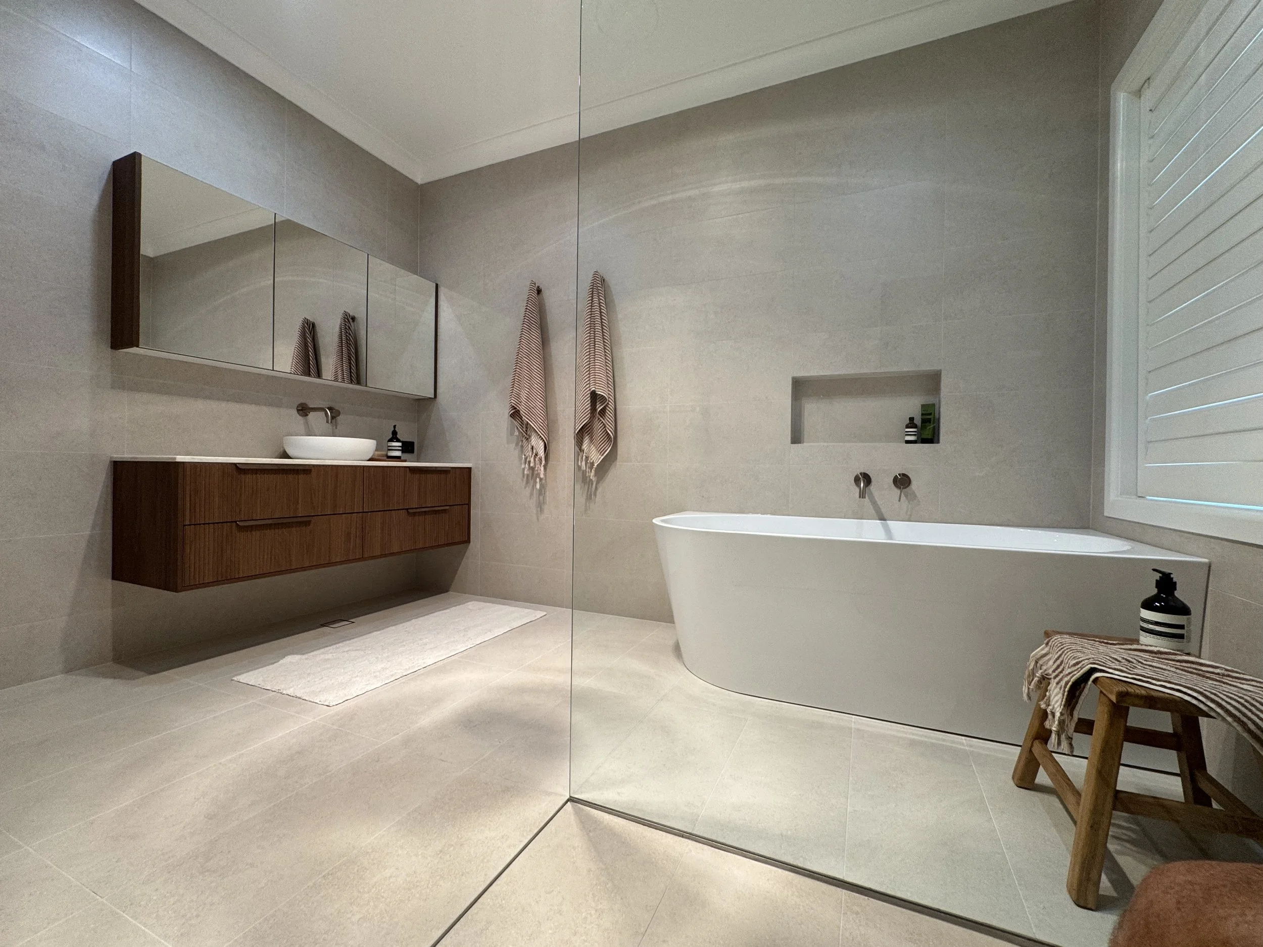 Modern bathroom featuring a white freestanding bathtub, a wooden vanity with a vessel sink, a large mirror, two striped towels hanging on the wall, a small bench with a striped towel, and a built-in shelf with bathroom essentials. There are natural l