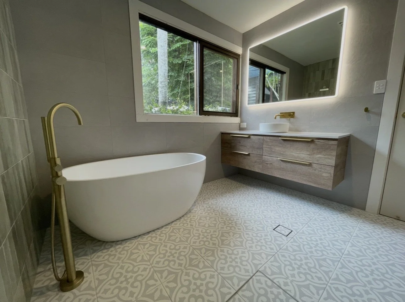 Modern bathroom with a freestanding bathtub, large window overlooking greenery, illuminated mirror, and a floating wooden vanity with a vessel sink and gold fixtures.