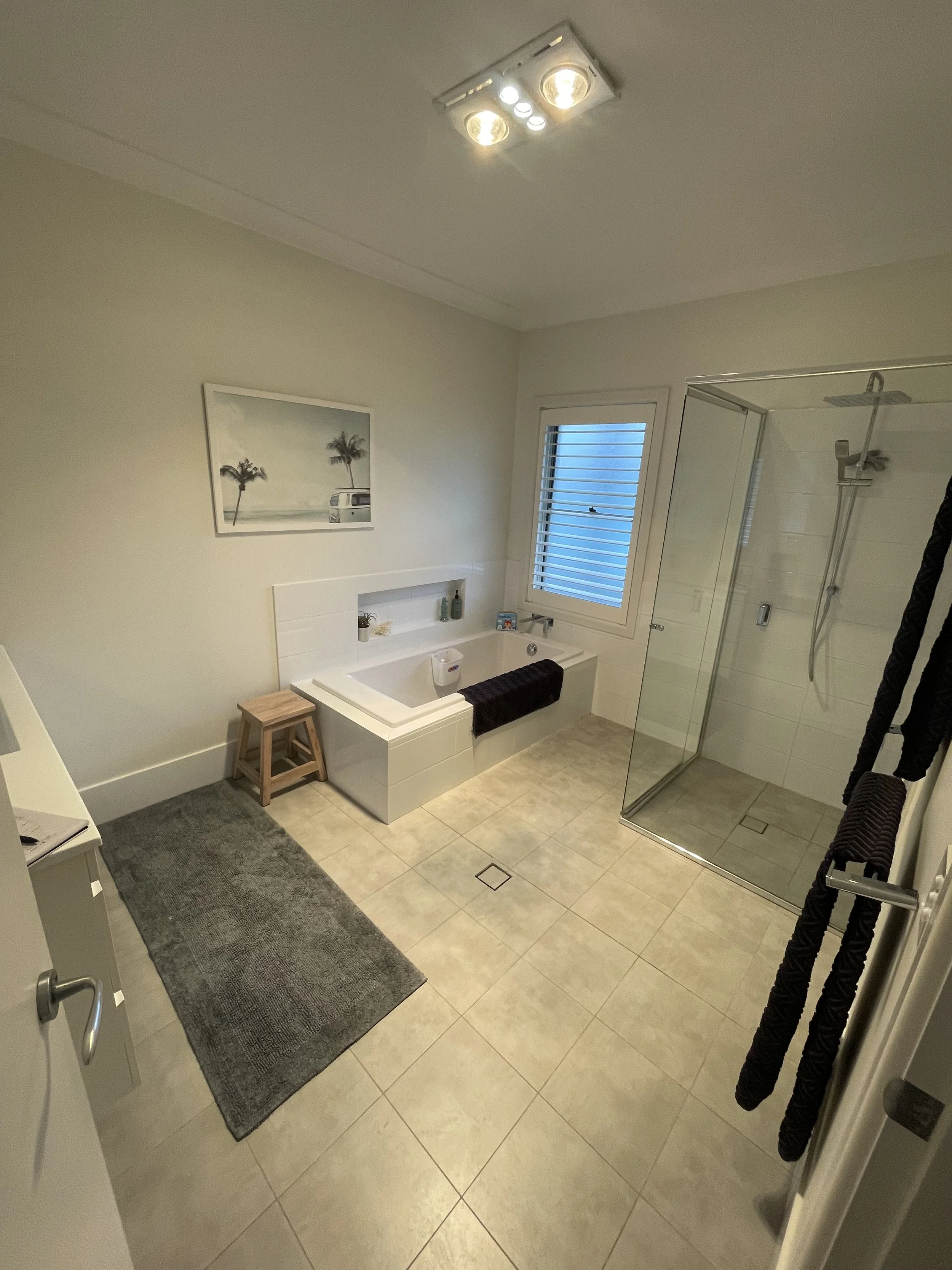 Bathroom with a bathtub below a window, a walk-in shower with glass doors, a towel rack with dark towels, a painting of palm trees and a van, a small wooden stool, a gray rug, and tiled floor.