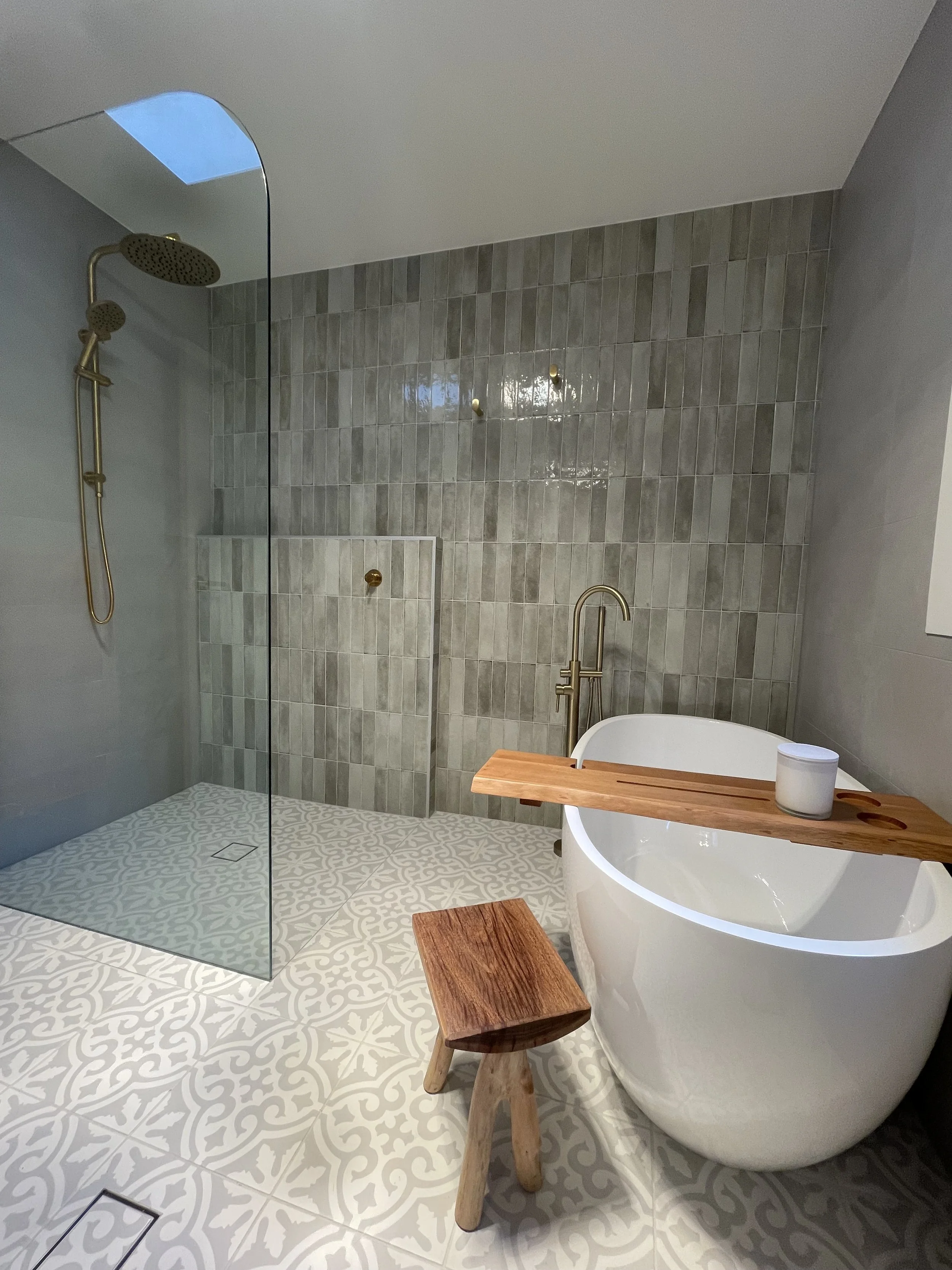 A modern bathroom featuring a walk-in shower with a brass showerhead and a glass partition, patterned floor tiles, a freestanding oval bathtub with a wooden tray and small stool, and a wall with gray tiles and brass hooks.