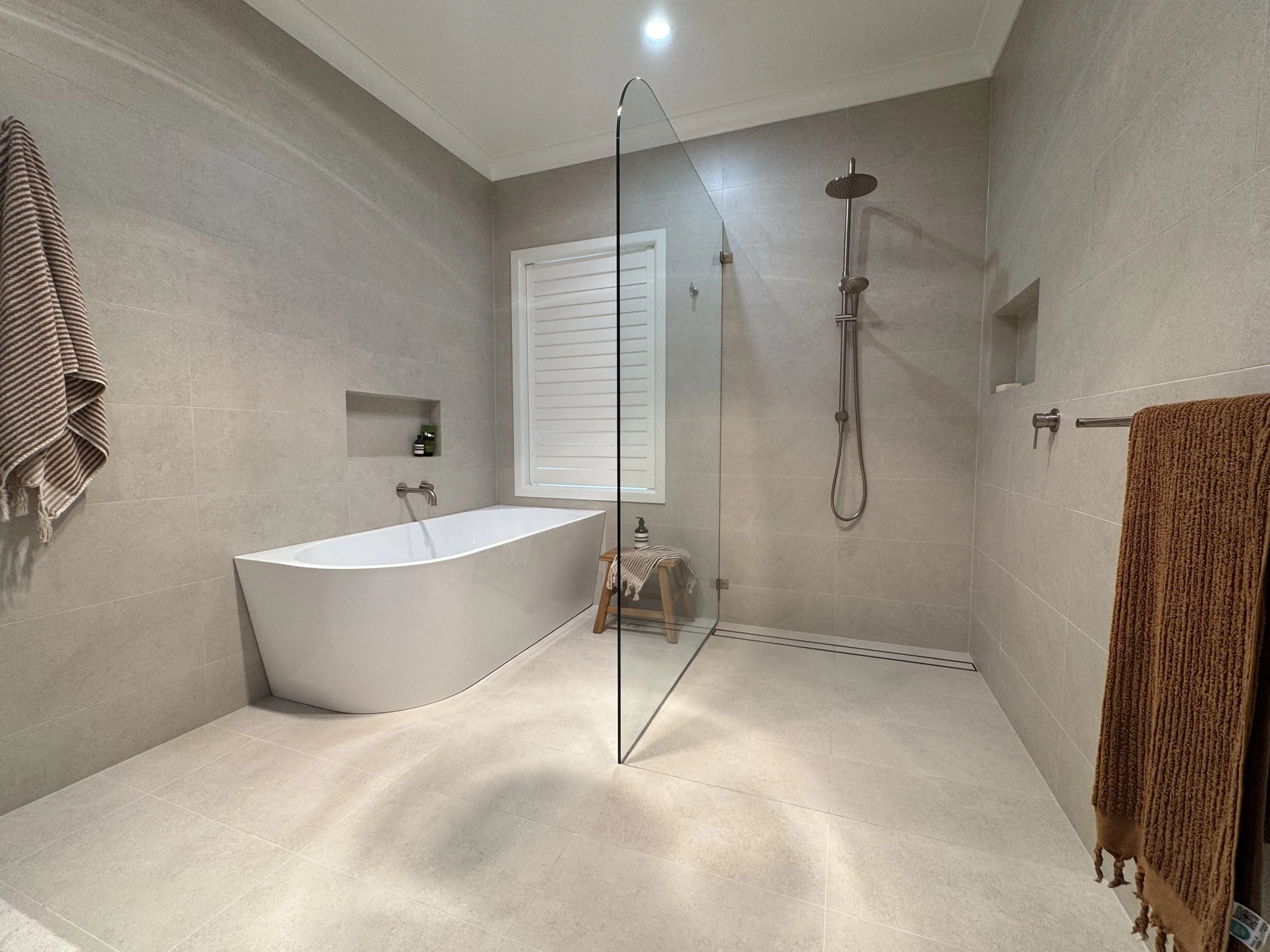 Modern bathroom with beige tiled walls and floor, a white freestanding bathtub under window blinds, a glass shower partition, a distinct showerhead, and towels hanging on racks.